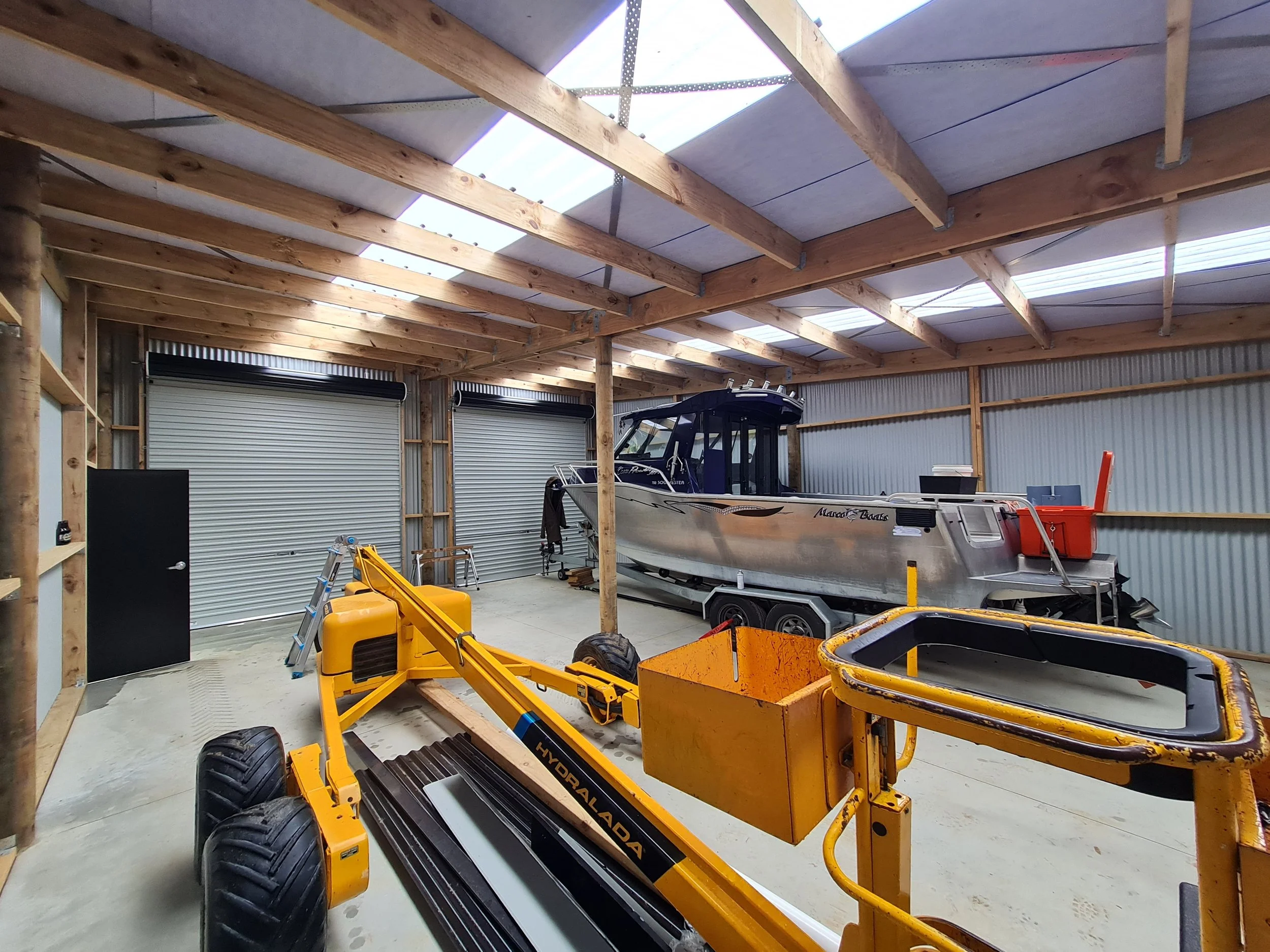A large metal boat parked inside a spacious garage with a wooden ceiling, along with yellow machinery, and storage items.