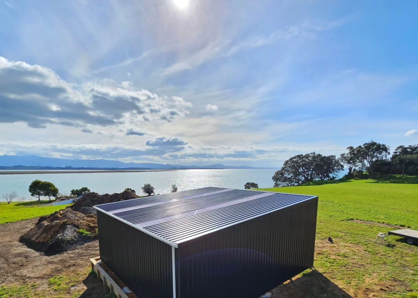 A black iron sand shed building with a corrugated metal roof situated and colour steel cladding on a grassy area near a body of water. The landscape features trees, rocks, and a large open sky with clouds and sunlight reflecting on the water.
