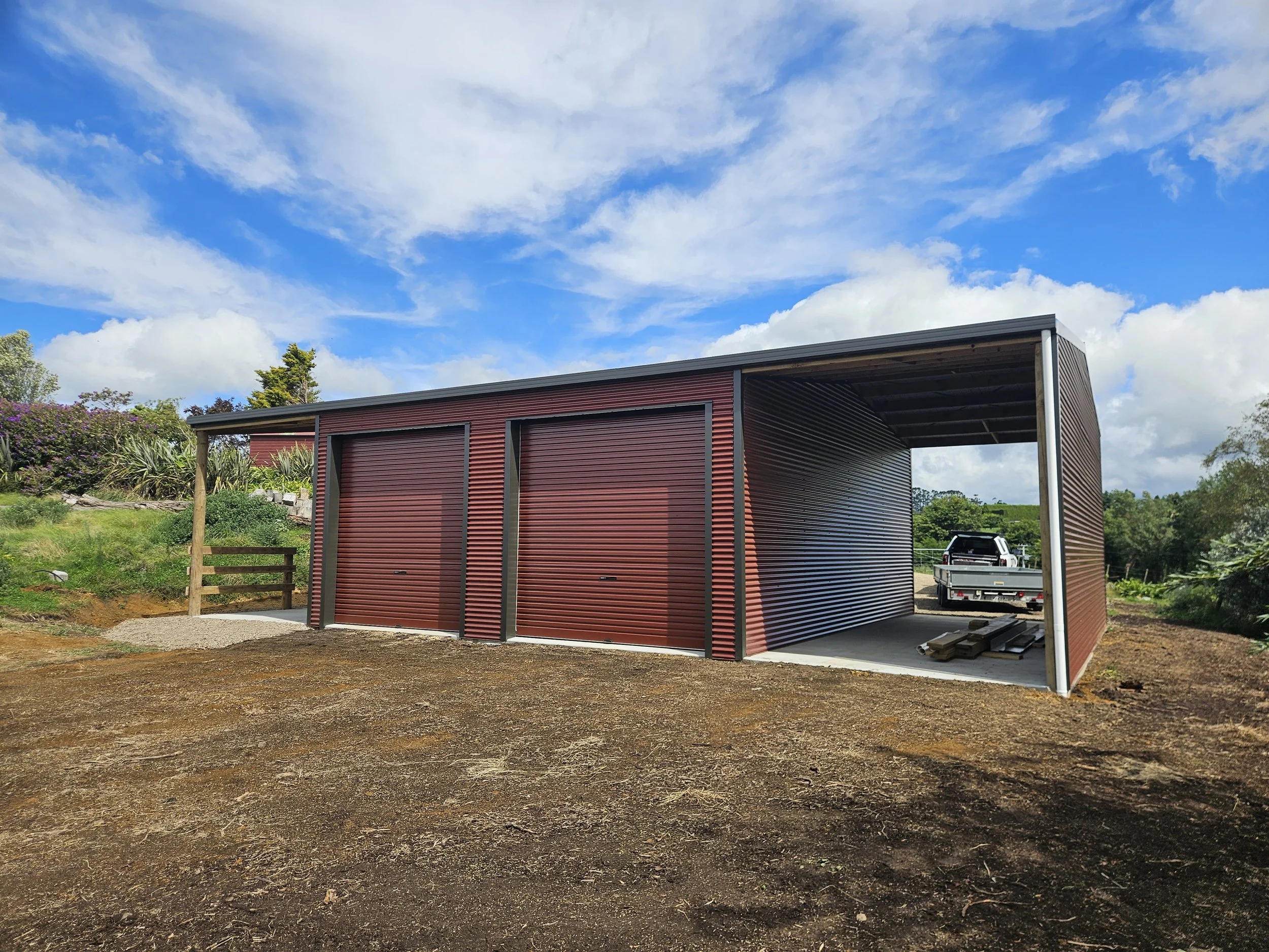 A rural modern cladded pole shed with pioneer red roller doors and an open side.  Horse Shed. Colour steel sides. A truck parked inside and stacked wood nearby, under a partly cloudy blue sky. In the Waihi Coromandel area