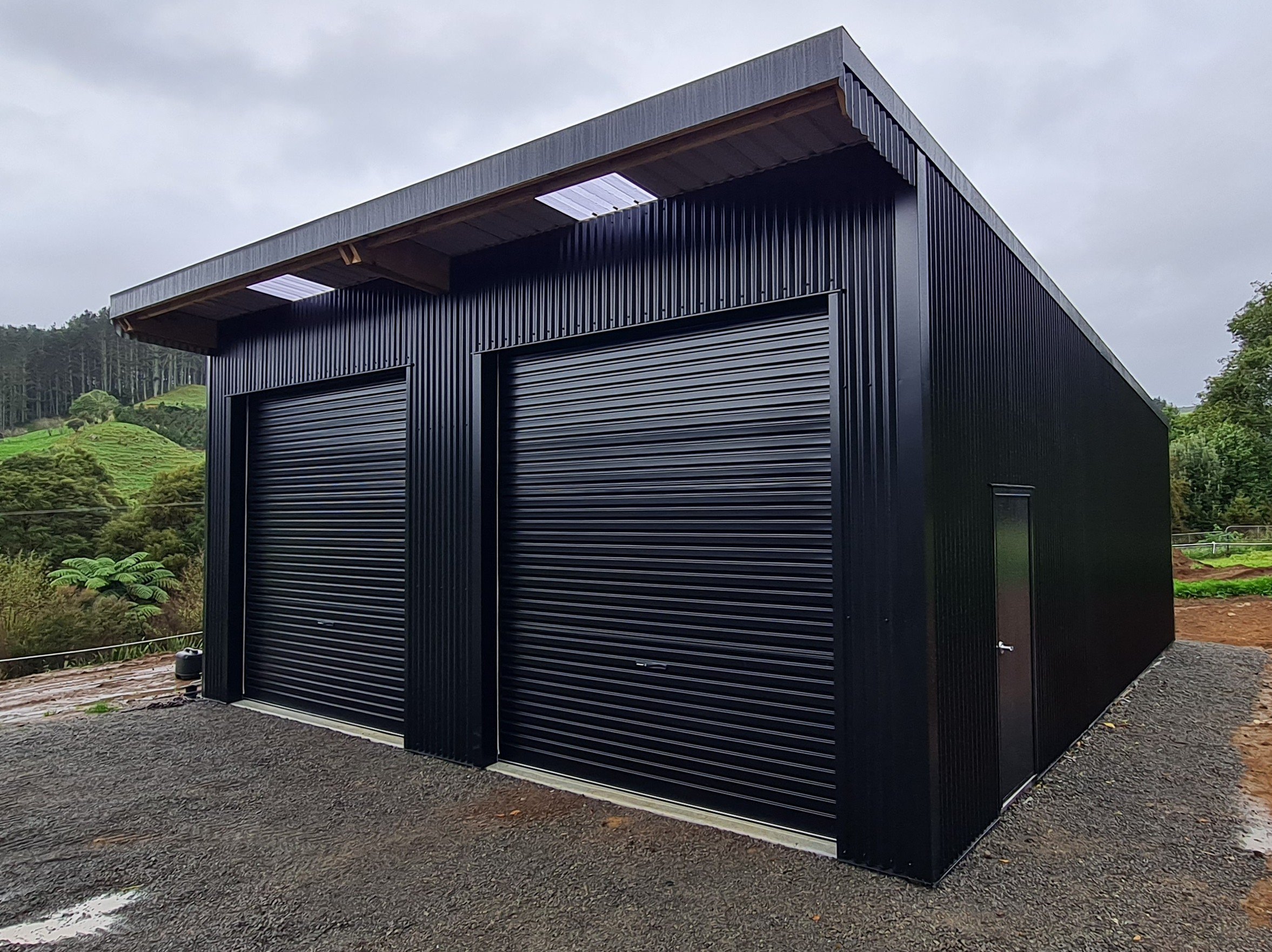 Black metal storage building with two roll-up doors and a small side door, set on a gravel driveway near green hilly landscape.