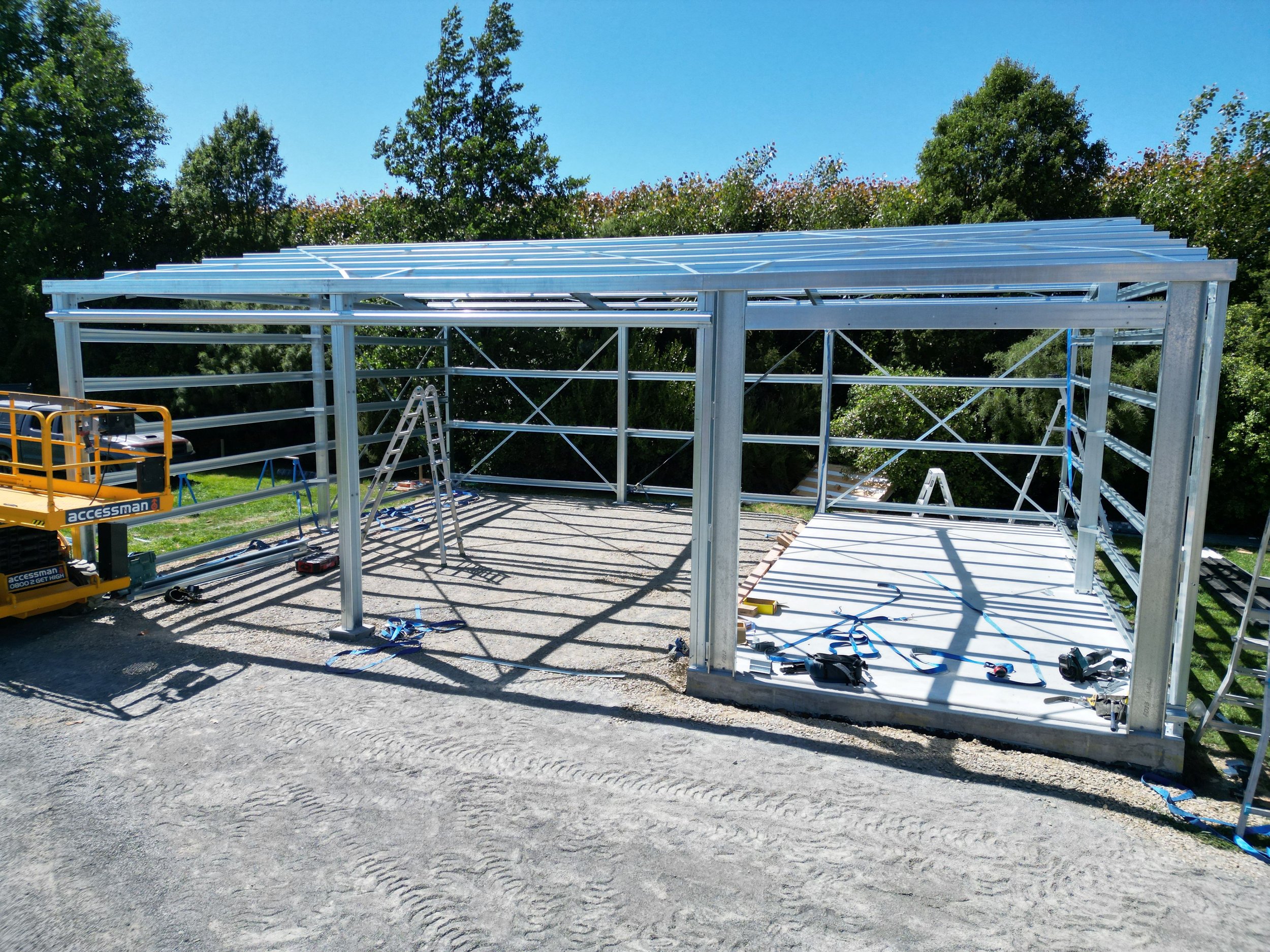 Steel frame of a building under construction with tools and equipment around, bright sunny day with trees in the background.