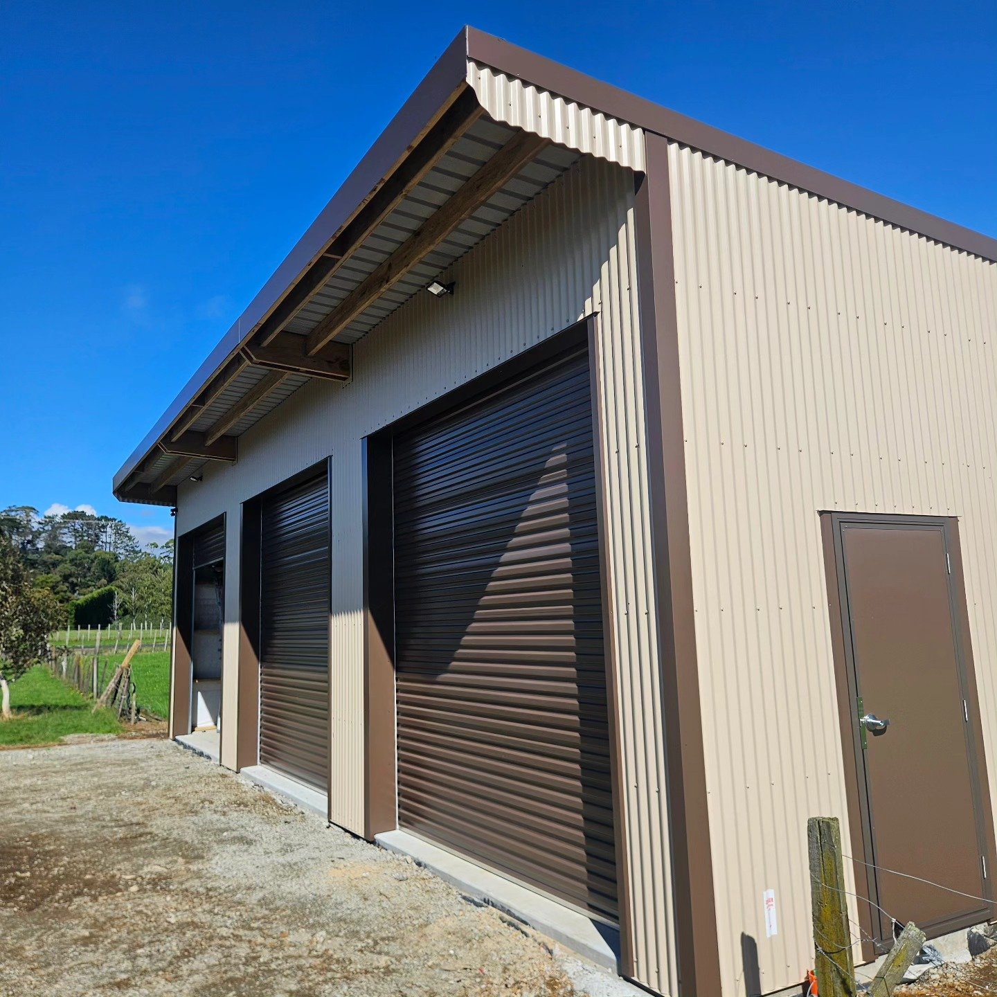 A metal building with three large roll-up garage doors and a side door, situated in a rural setting under a clear blue sky.