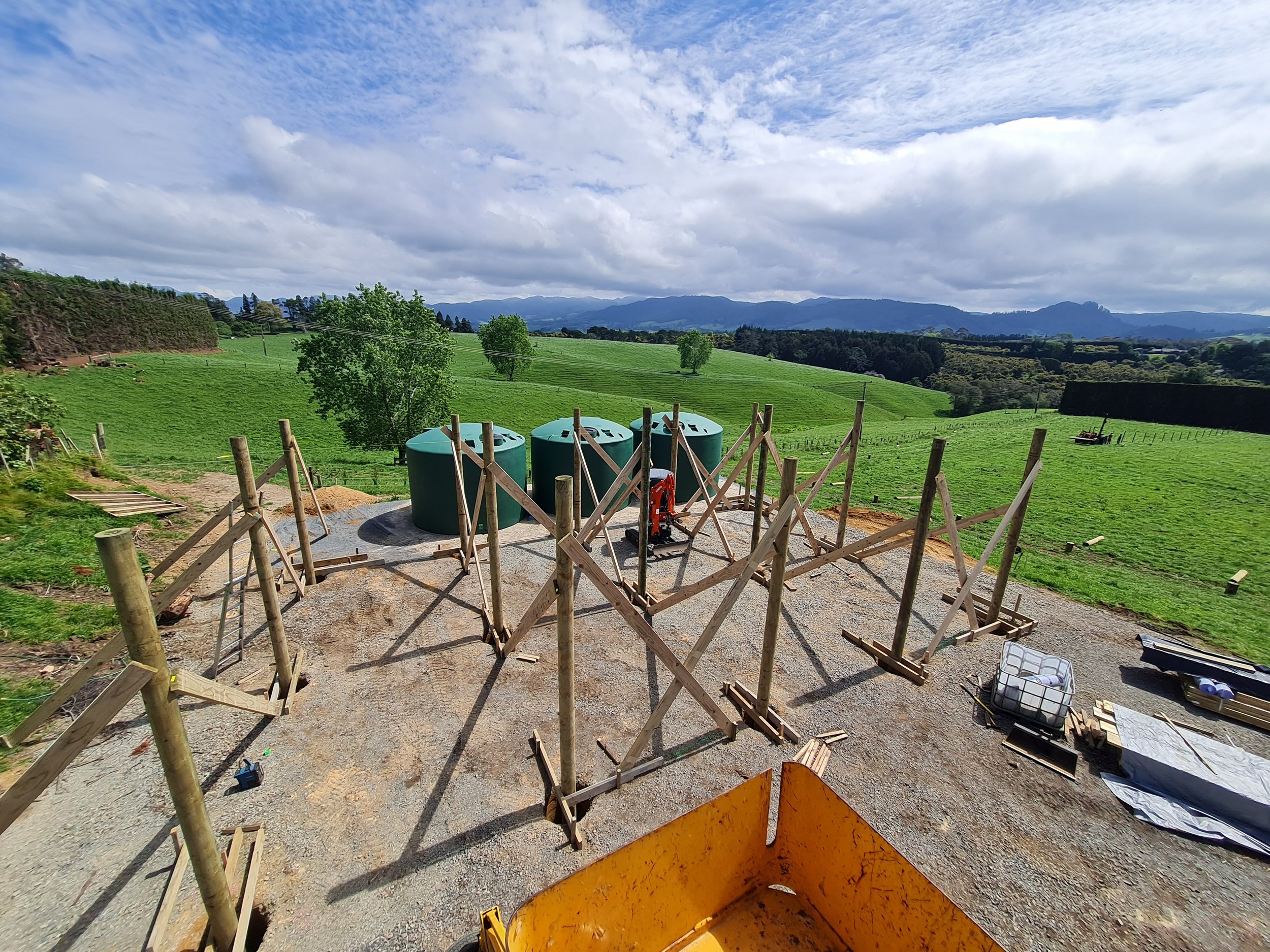 Construction site with wooden framework and large water tanks in a green rural landscape under a partly cloudy sky.