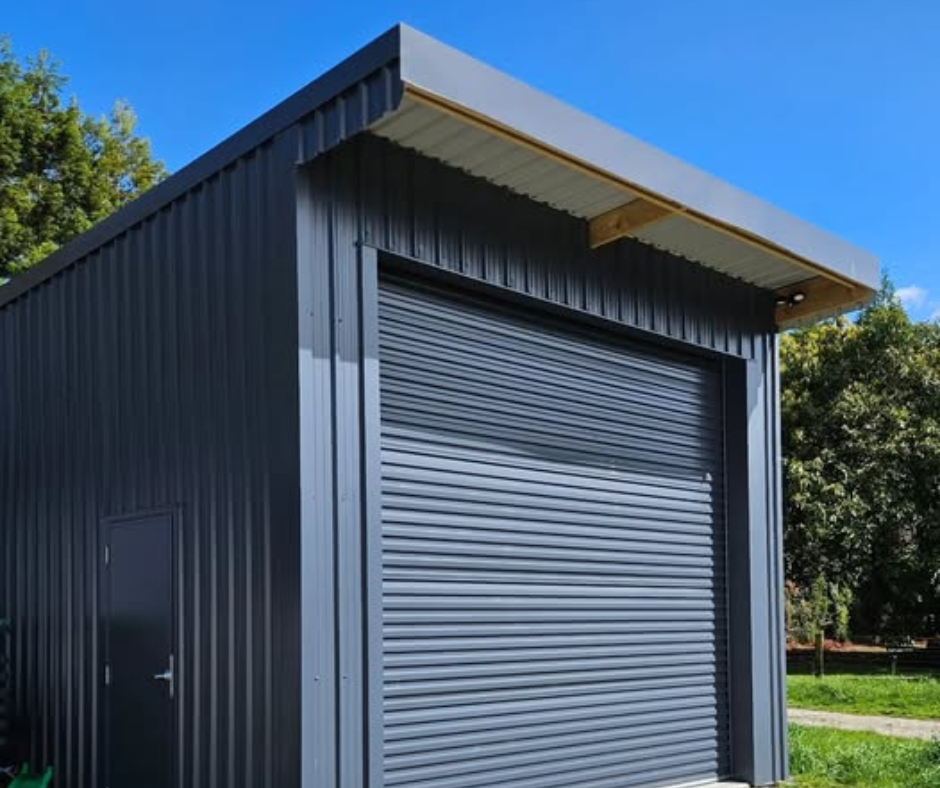 A small, dark gray metal building with a roll-up garage door and a side door, surrounded by green trees and grass under a blue sky.