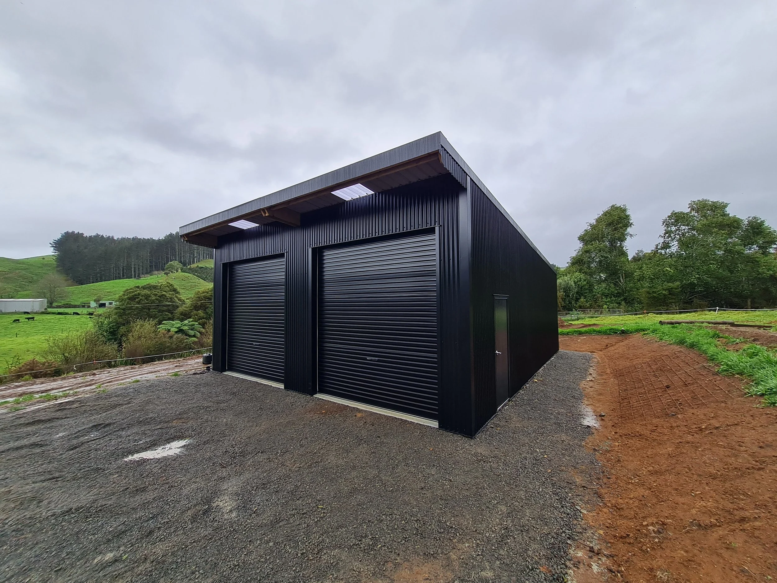 A black metal storage building with two large rolling doors and a small side door, situated in a rural area with green fields, trees, and cloudy sky in the background.