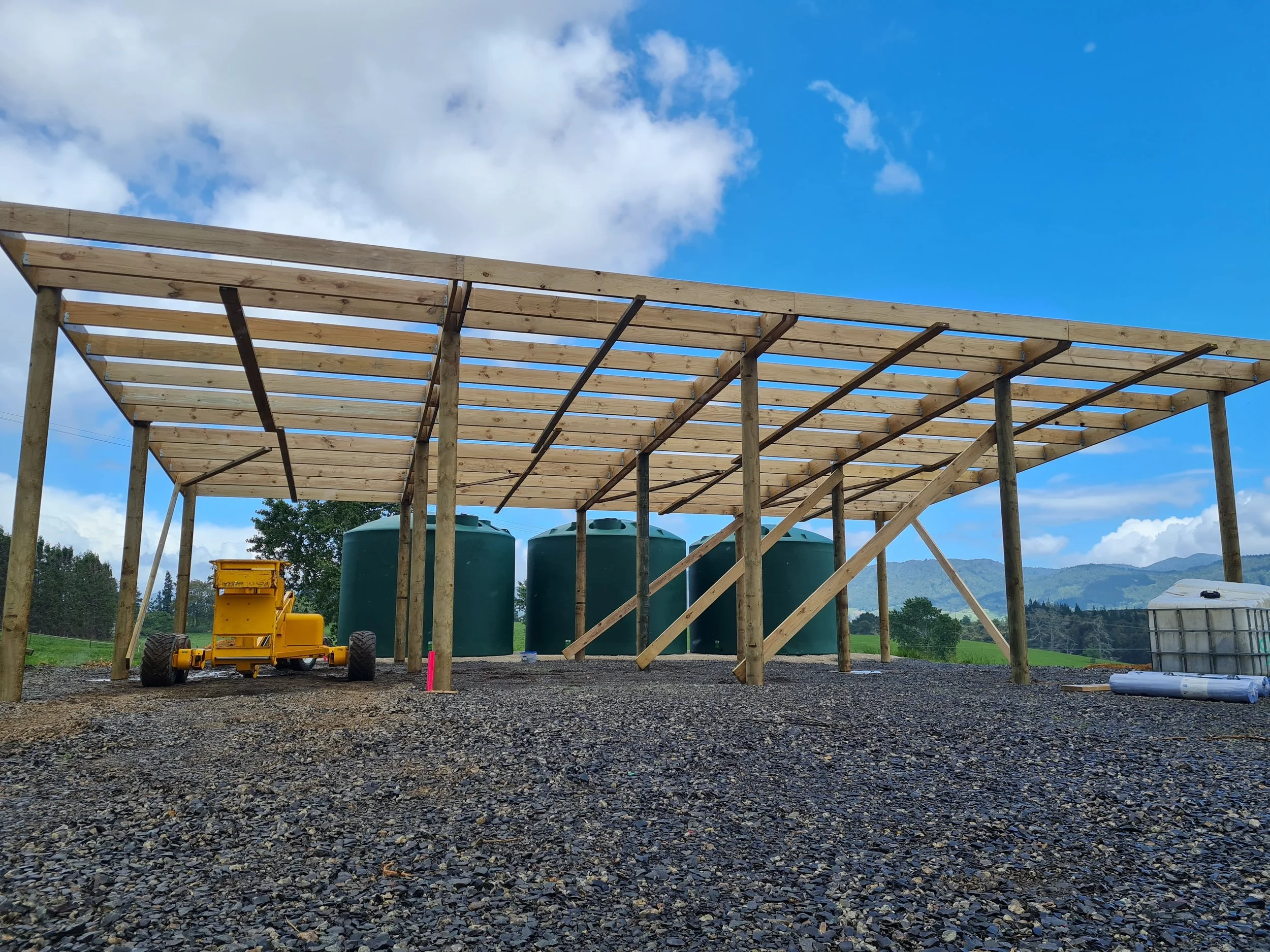Construction site with a large industrial  wooden frame pole shed being constructed, three green water tanks, a yellow machine, and a cloudy sky with blue background.