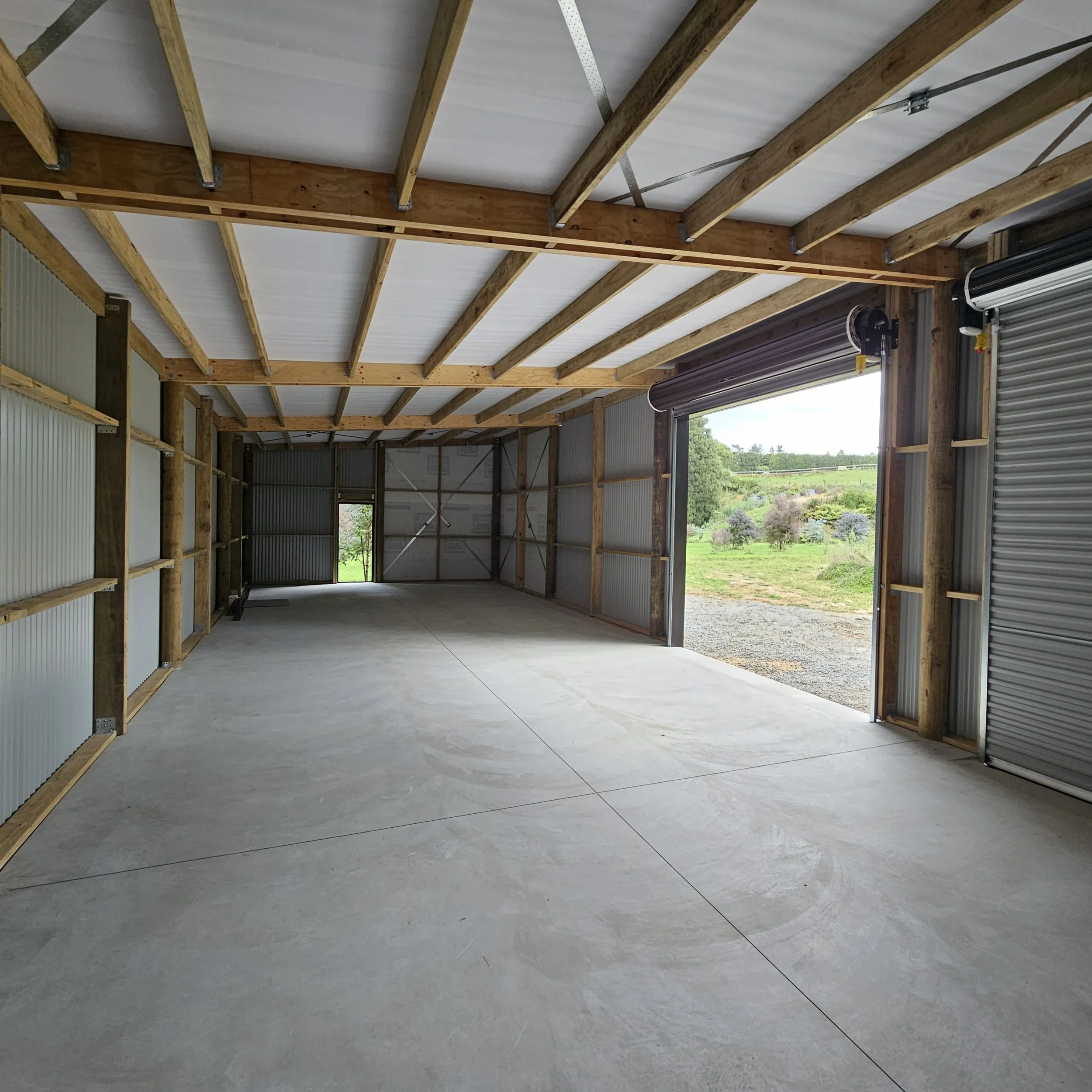 Empty garage with concrete floor, wooden framing, and partly open roll-up door revealing outdoor greenery.