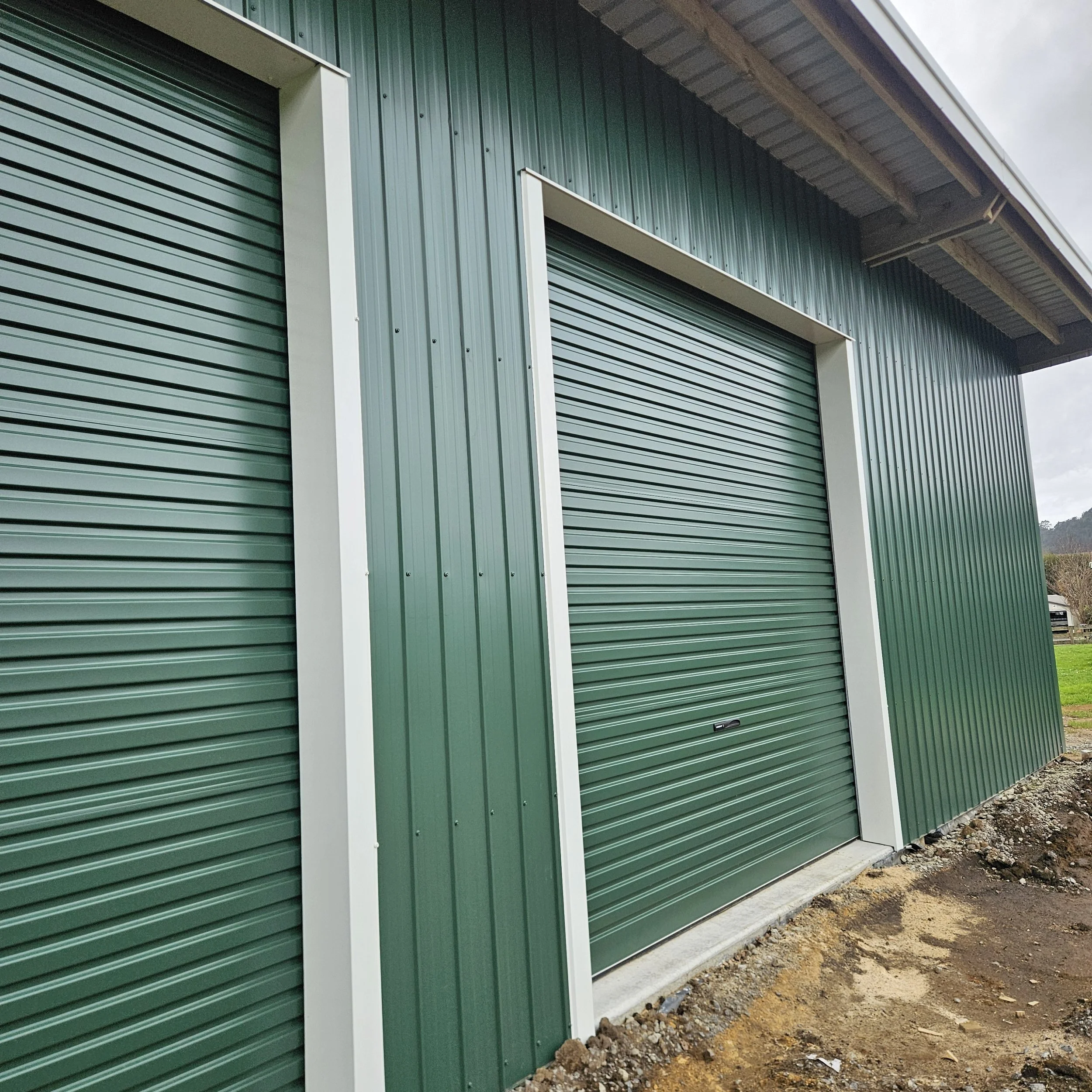 Colour steel green cladded building with roller doors, one partially visible on the left and the other large door in the center.  On a orchard in Athenree. 