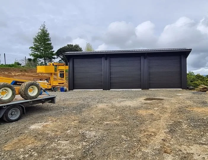 Iron sand building or shed with a sloped roof, set on a gravel lot. 3 bay roller doors and personal access door. To the left, there is a yellow piece of construction equipment on a flatbed trailer, with large tires. KatiKati. 