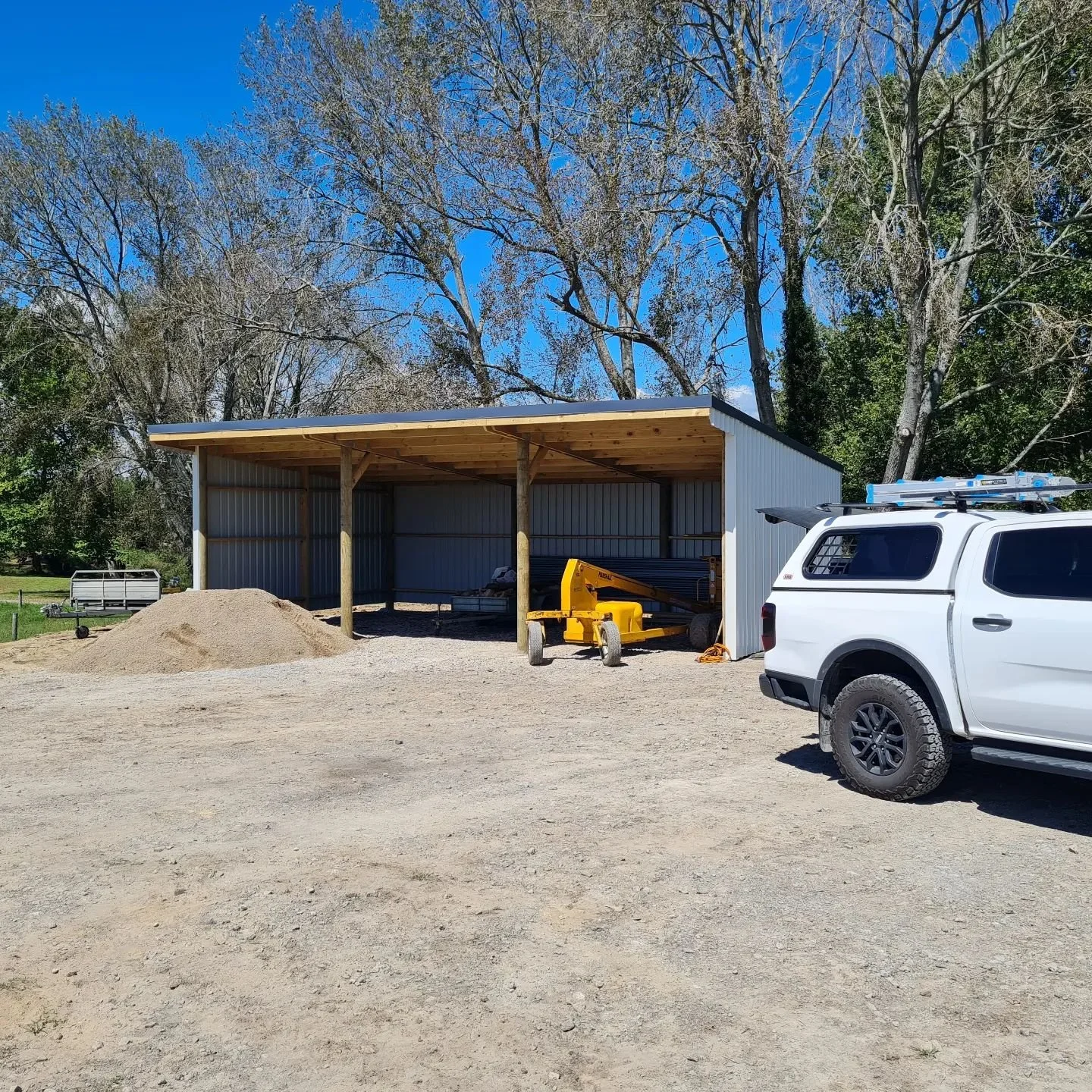 A partially enclosed metal 3 bay shed with an iron roof, with gravel and construction equipment in front, and a white pickup truck parked nearby on a gravel surface, with trees in the background under a clear blue sky. Waihi Athenree 