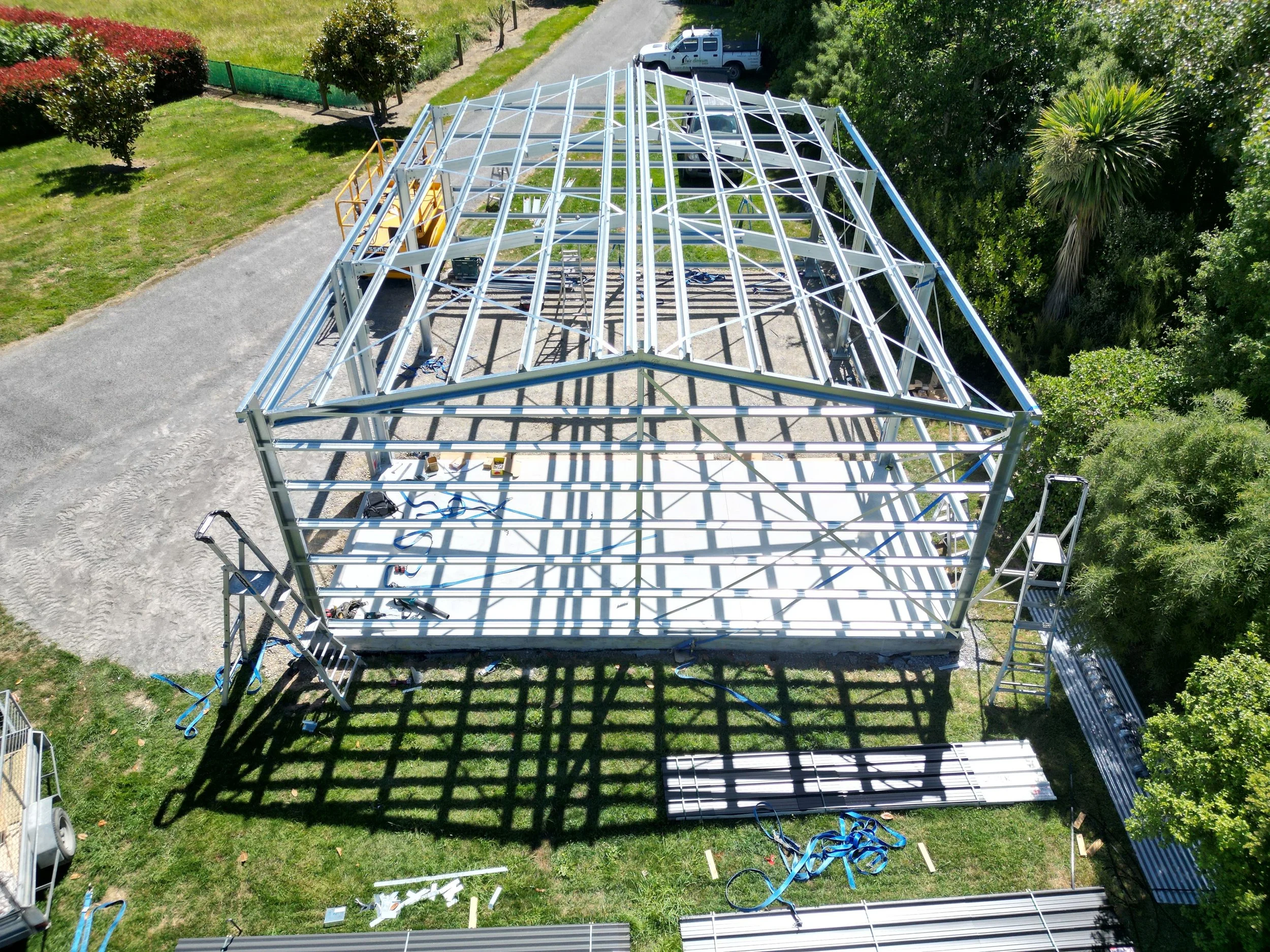A  steel frame shed under construction with a metal frame structure, scaffolding, and ladders, surrounded by trees and grassy areas.