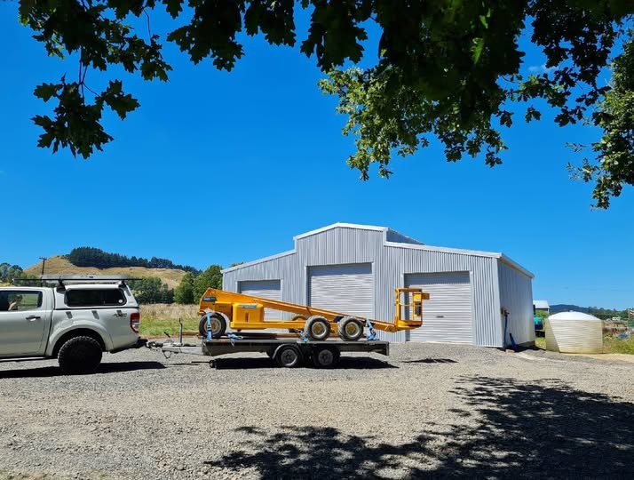 A white utility American style barm with large garage doors, surrounded by a gravel area. In front, a silver truck is towing a trailer with a yellow piece of equipment. There are trees above and hills in the background. This is in rural Waihi