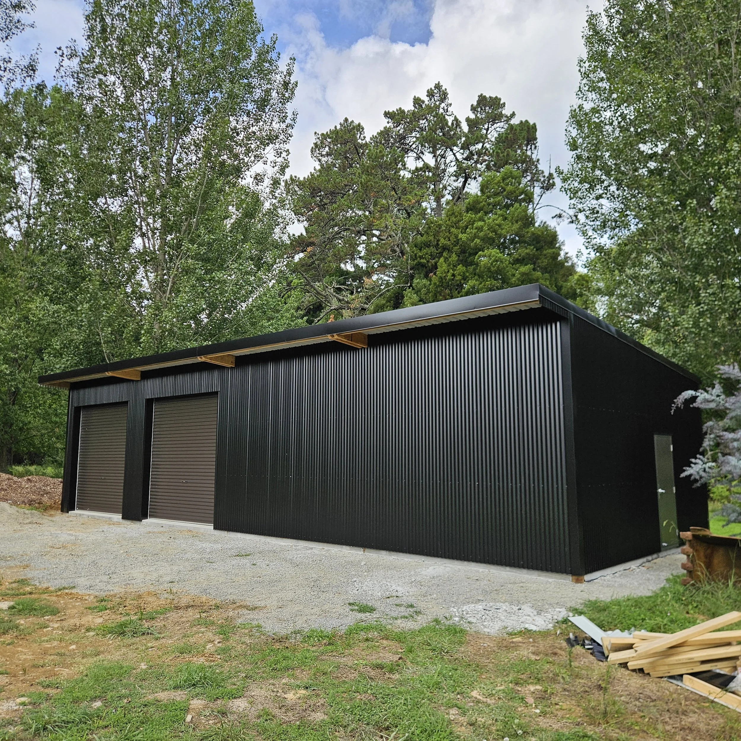 Black metal garage with two roll-up doors and a side entrance, situated on a gravel pad with trees in the background under a partly cloudy sky.