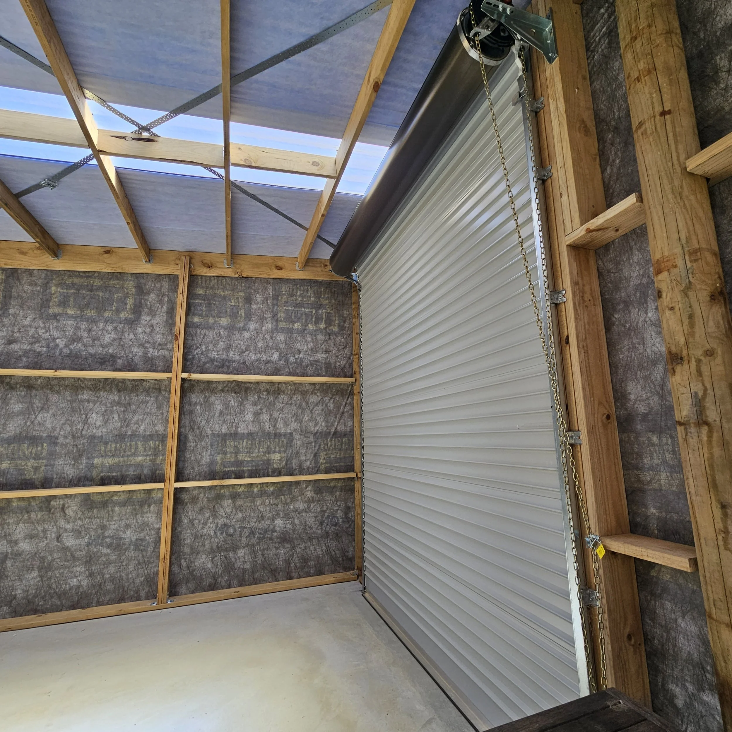 The interior of a garage under construction, showing wooden framing, insulation, and a partially installed roll-up metal garage door.