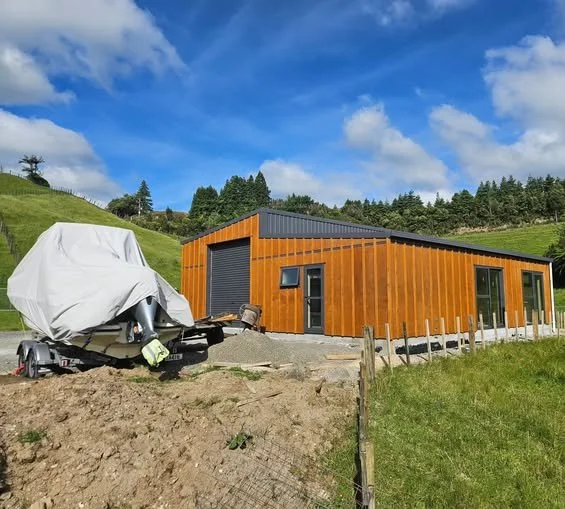 Modern Tan and Gun metal blue pole shed building. Situated on grassy hillside with a boat covered by a tarp nearby and a partly cloudy sky in the background. Waihi. 