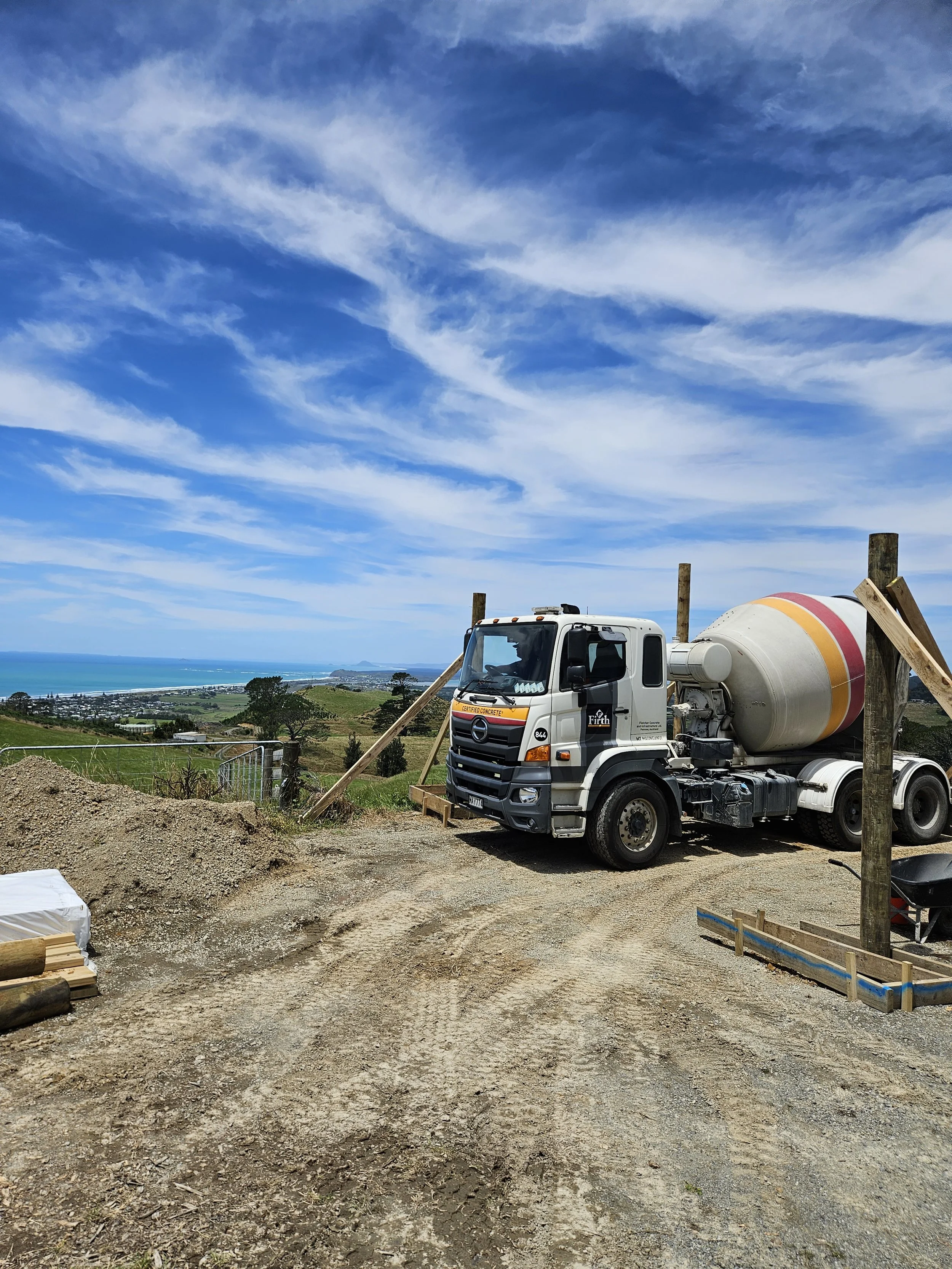 Construction site with a cement mixer truck on a dirt road overlooking a scenic ocean view under a partly cloudy sky.