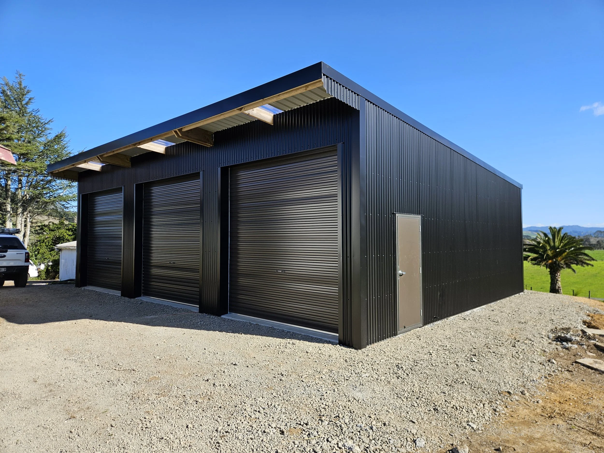 A black colour steel shed with three roll-up garage doors and a side door on a farm. Set on a gravel lot with a green field and palm trees in the background. Iron sand colour.  Area of Katikati Waihi. 