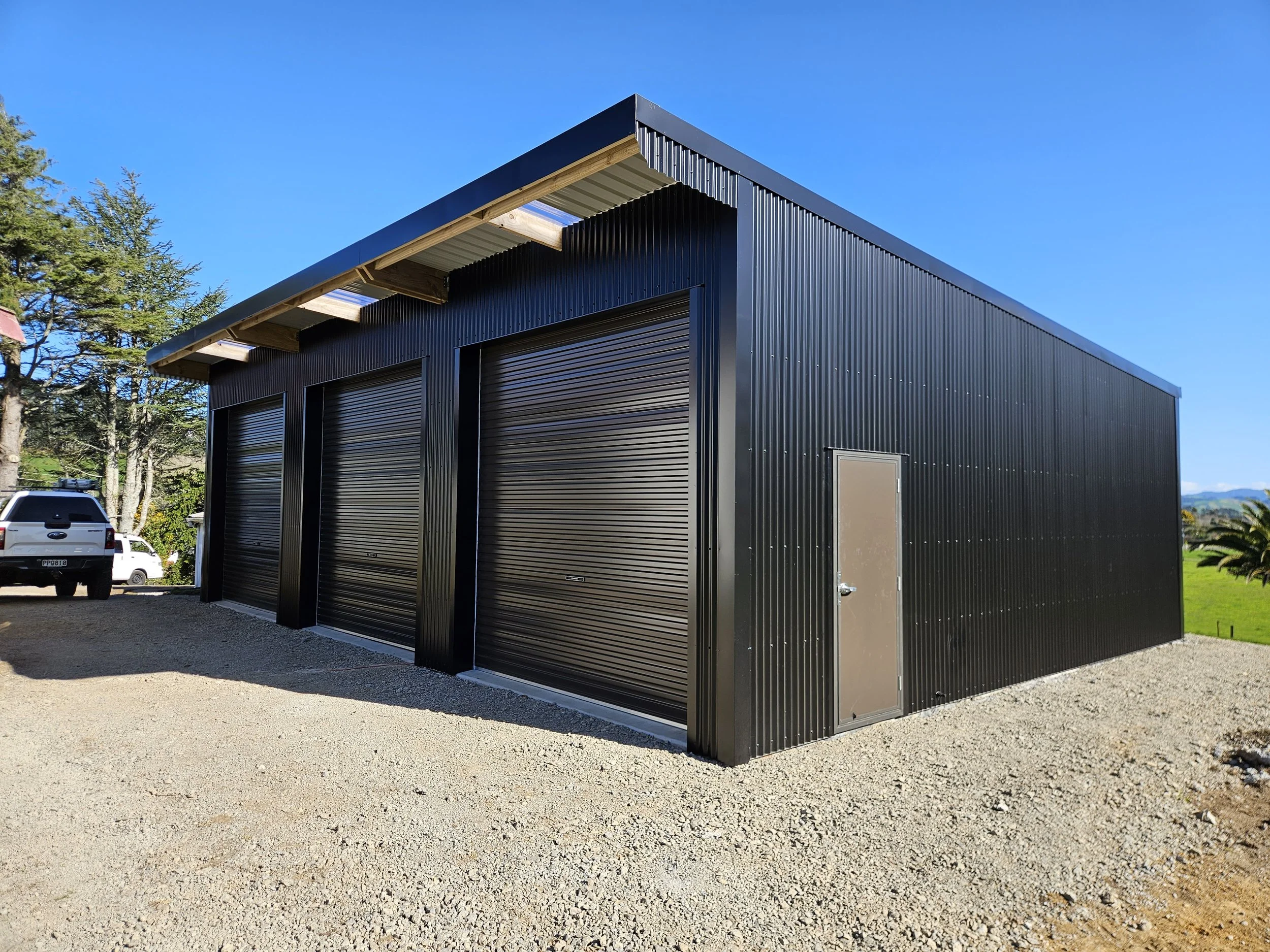 Black metal building with three garage doors and one side door, set on a gravel lot with trees and clear blue sky in the background.