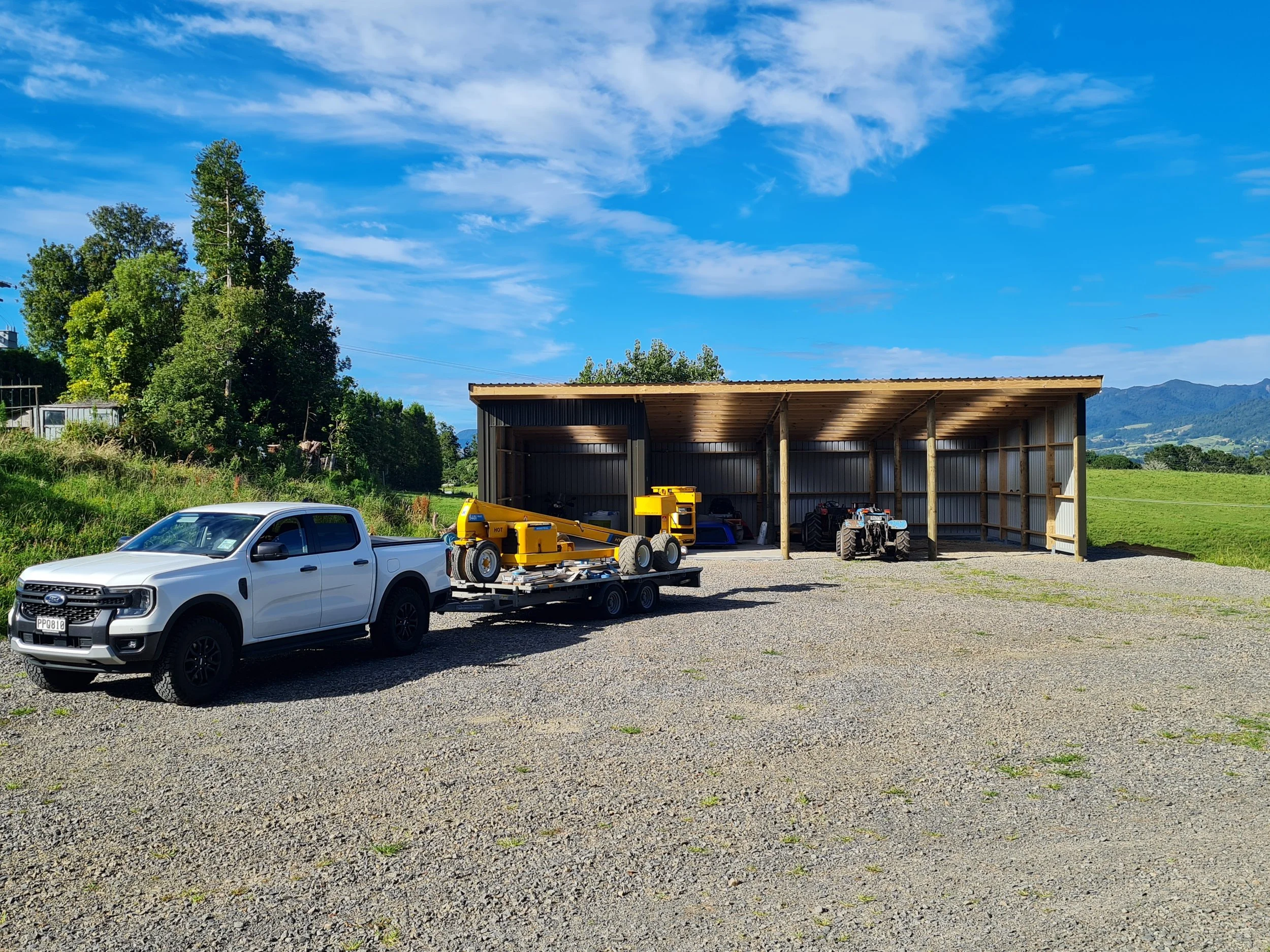 A white pickup truck parked on gravel with a trailer carrying yellow construction equipment, and a wooden shed with various machinery inside, in a rural area with green fields, trees, and mountains in the background.
