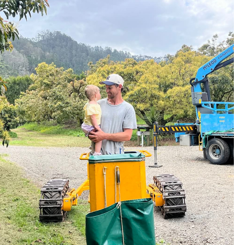 A man holding a young girl in a yellow shirt and purple shorts outdoors in a park or garden with trees and mountains in the background. There is industrial equipment, including a yellow tracked vehicle and a blue crane, nearby.