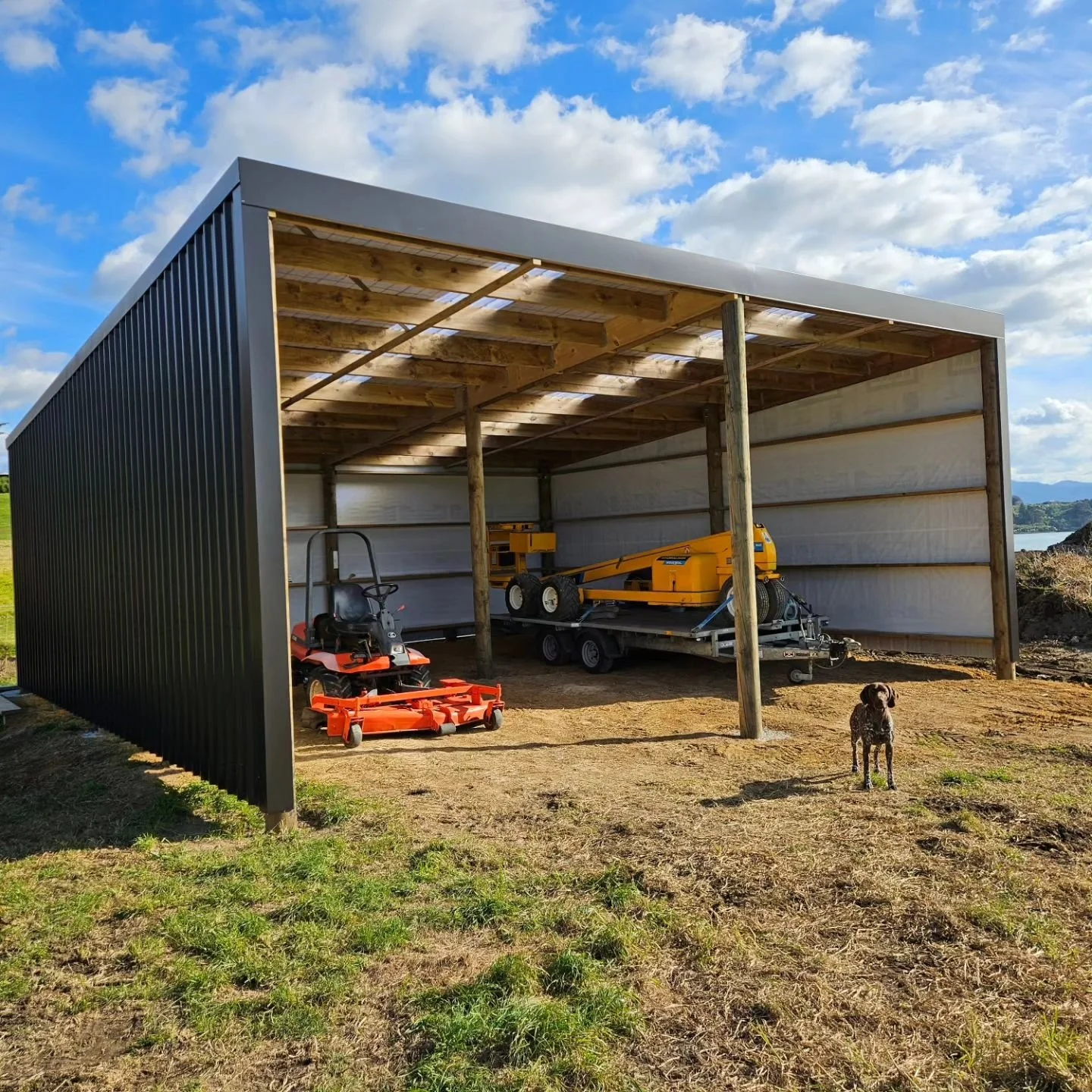 A rural pole shed with machinery stored and construction materials inside, including a ride on lawn mower, a yellow lift, and a small dog standing on the dirt outside. Consent free