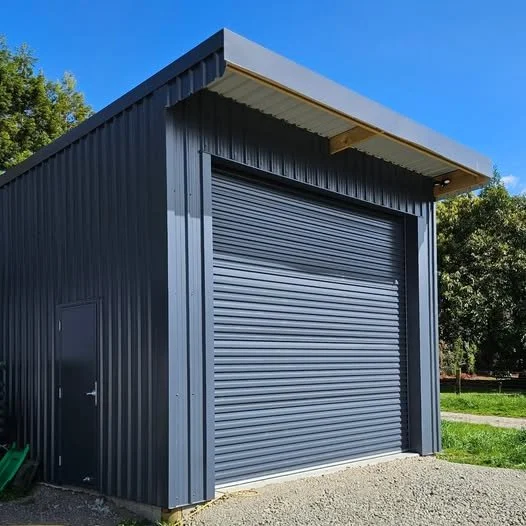 A small industrial blue metal building with a roll-up garage door and a side door, set outdoors with trees and a clear blue sky in the background.