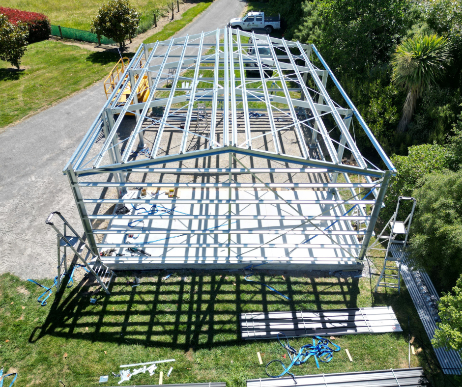 A metal framework for a building under construction, with ladders and tools on the ground, surrounded by greenery and a gravel path, and a white truck parked nearby.