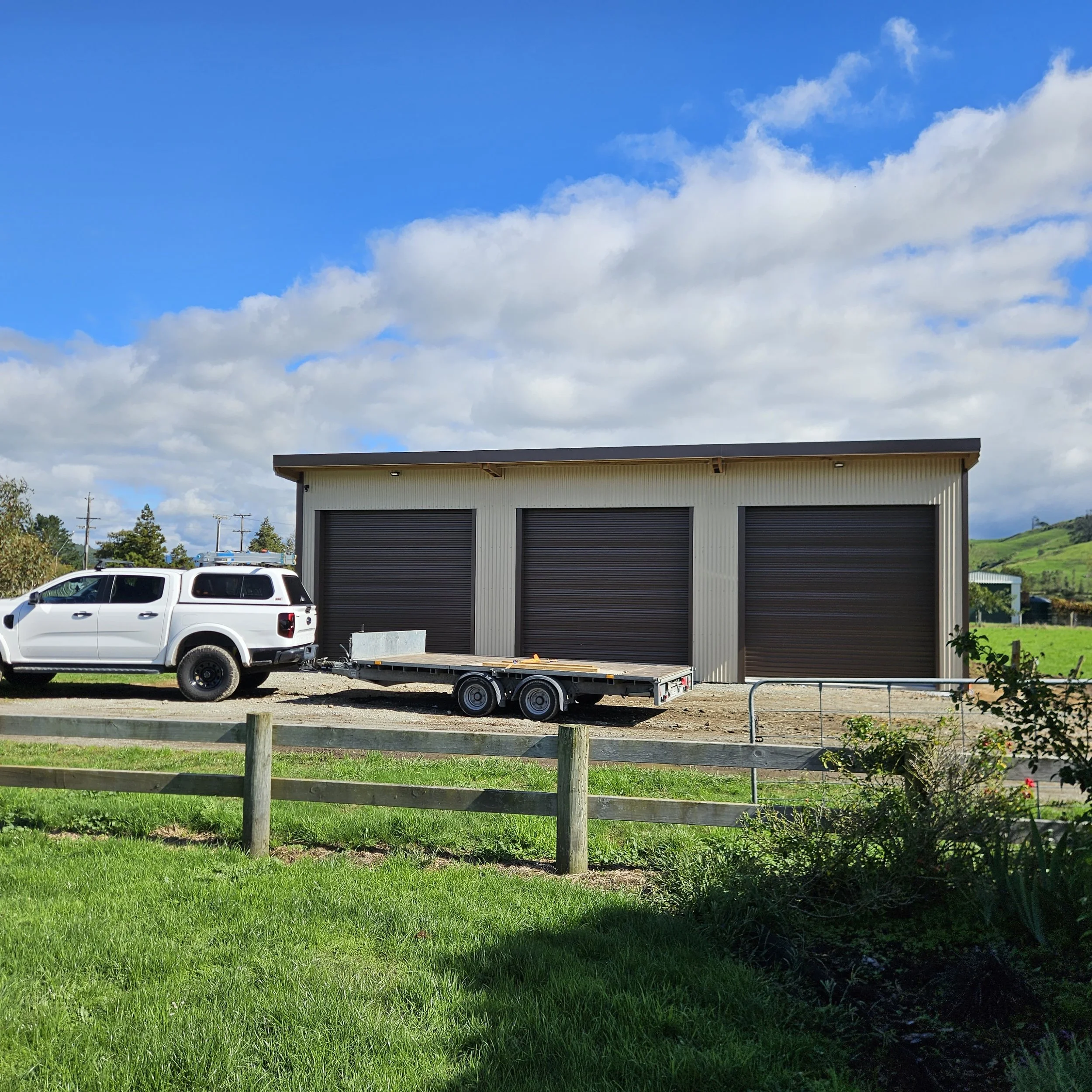 A large pole shed with three roller garage doors, a white pickup truck parked nearby towing a flatbed trailer, and green fields with rolling hills in the background under a partly cloudy sky. Waihi.