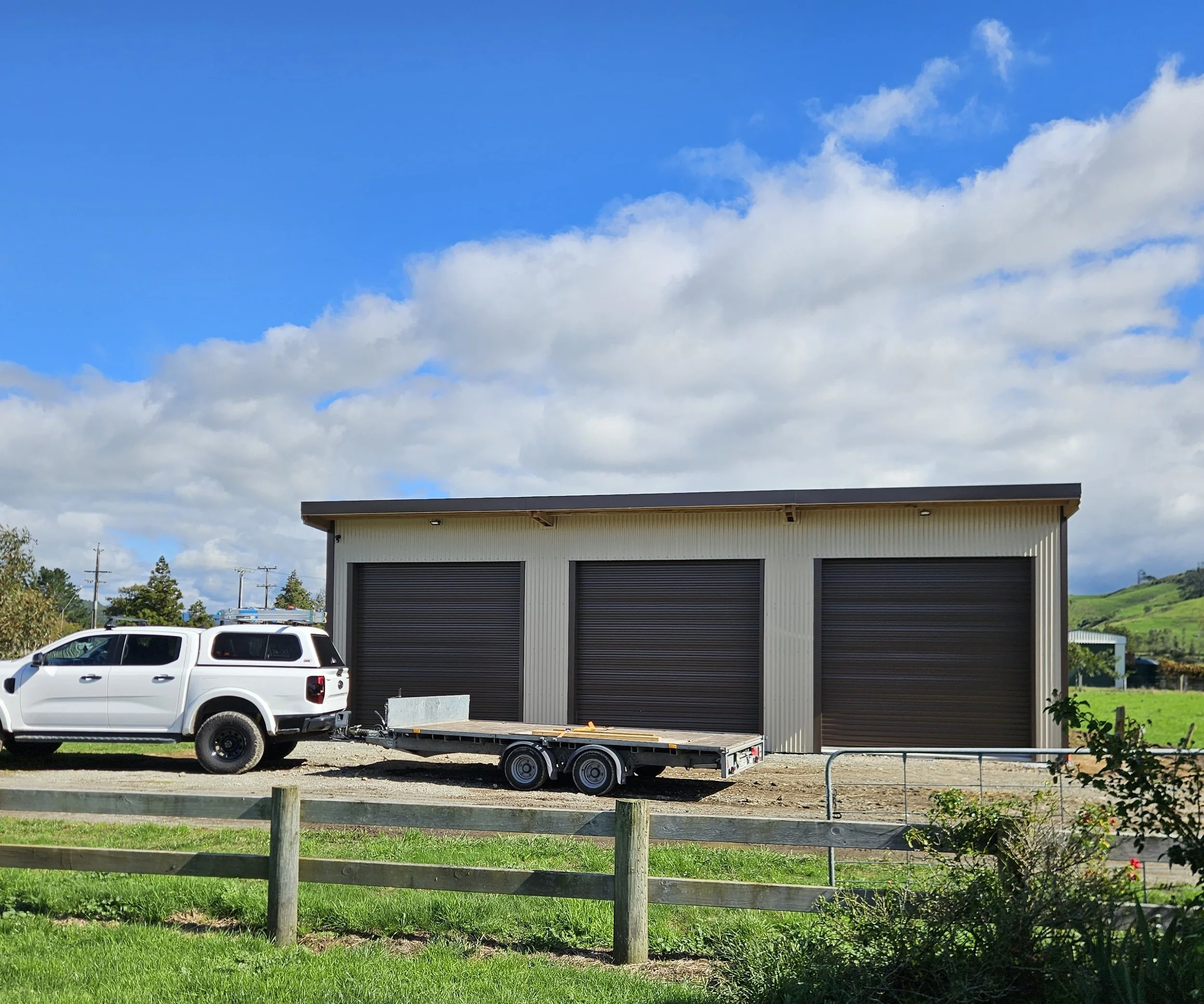 A white pickup truck with a trailer parked in front of a storage garage with three black roll-up doors, set against a partly cloudy sky and green hills in the background.