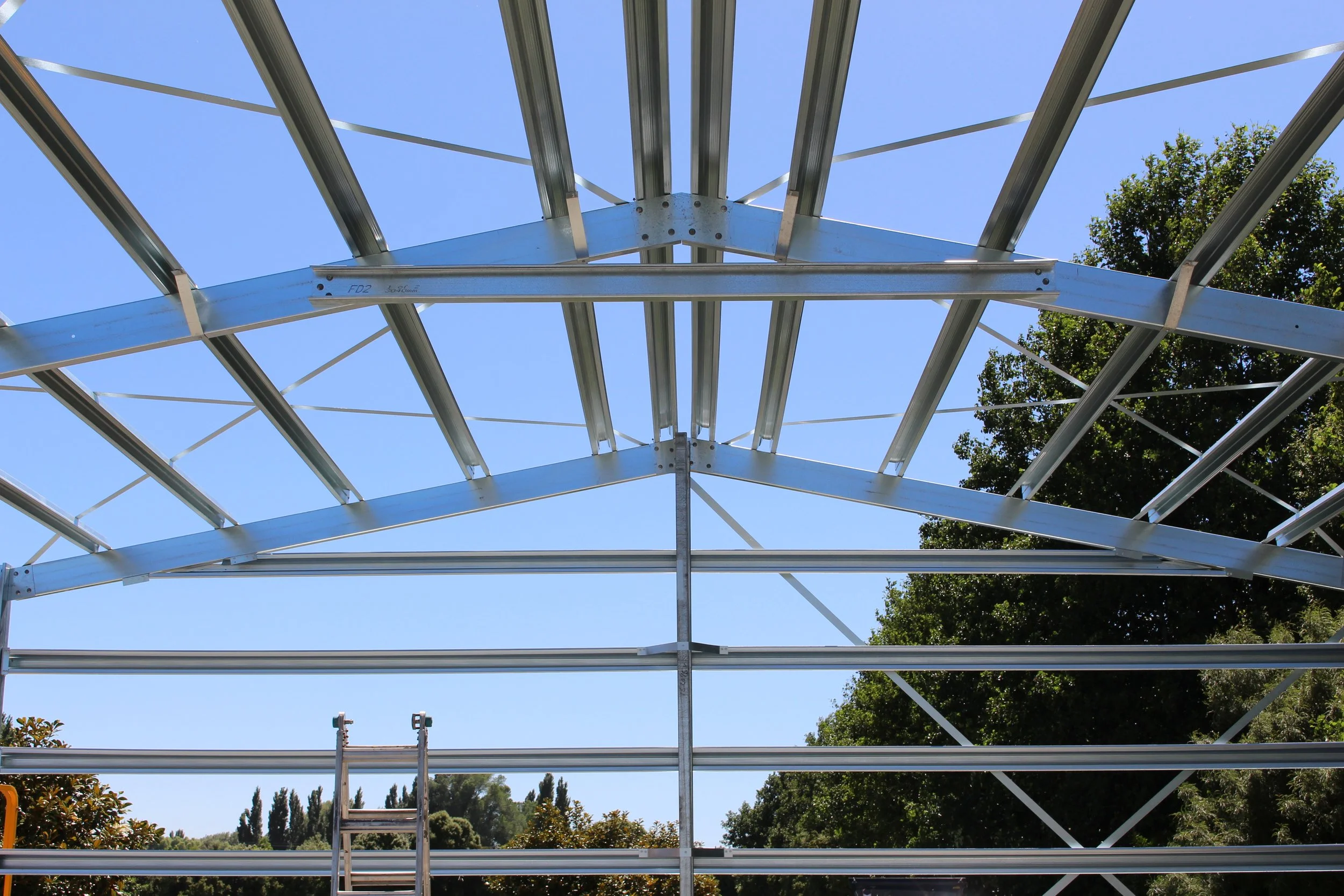 Looking up at a metal framework of a steel frame shed under construction with a ladder visible, set against a clear blue sky and surrounded by trees.  Waihi 