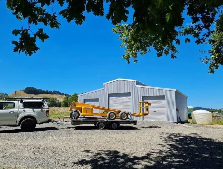 A metal building with two large garage doors, parked on a gravel lot with a trailer carrying a yellow piece of equipment, and a white pickup truck, under a tree with a clear blue sky.