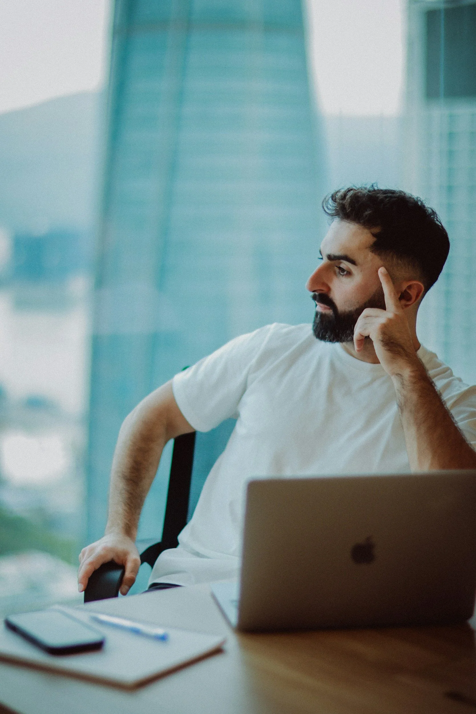 A man with dark hair and beard, wearing a white t-shirt, sitting at a desk with a laptop in a modern office space, with large windows in the background.