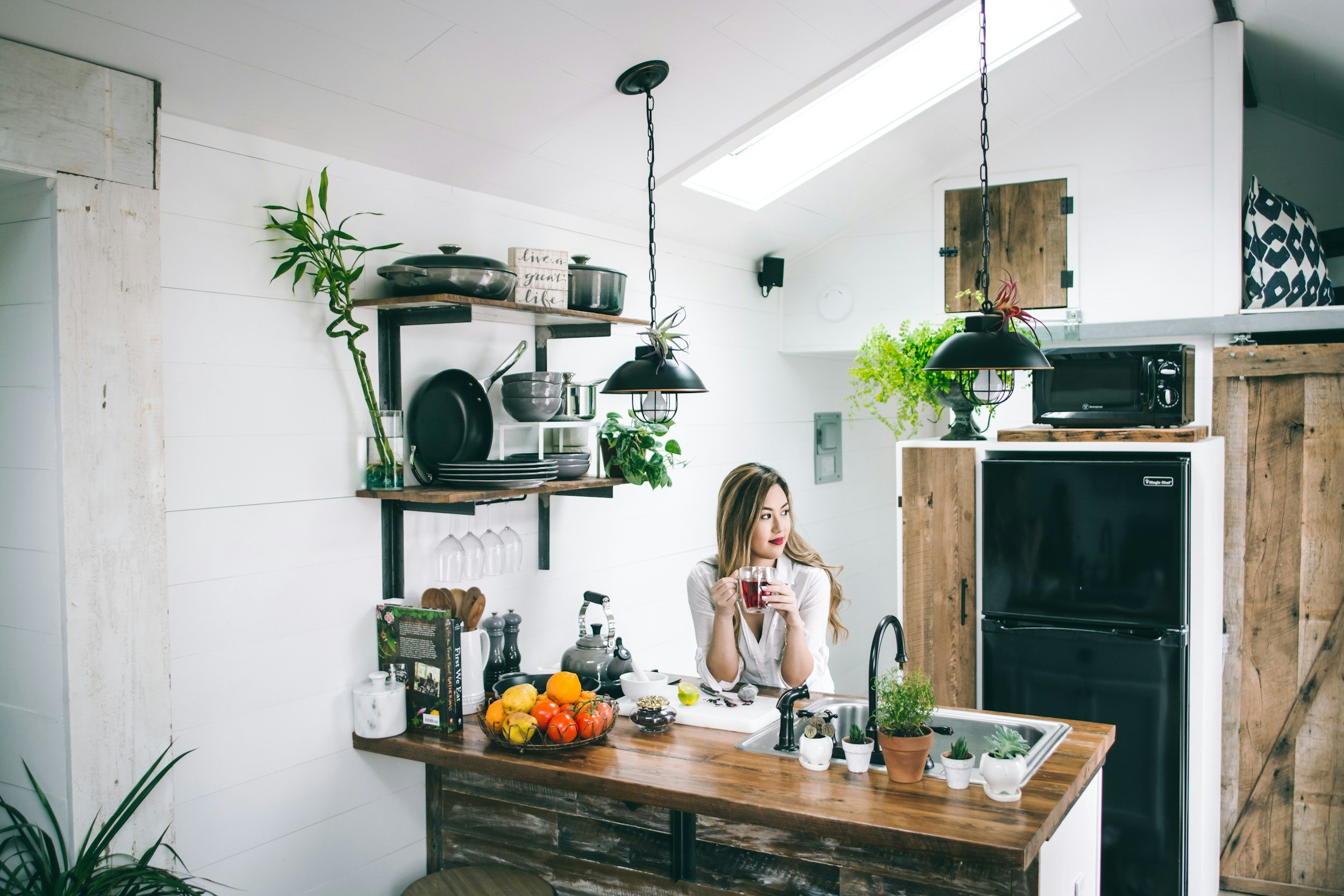 Woman sitting at a kitchen counter holding a glass of red beverage with a bowl of vegetables and various kitchen items on the counter, surrounded by plants and kitchen shelves with cookware.