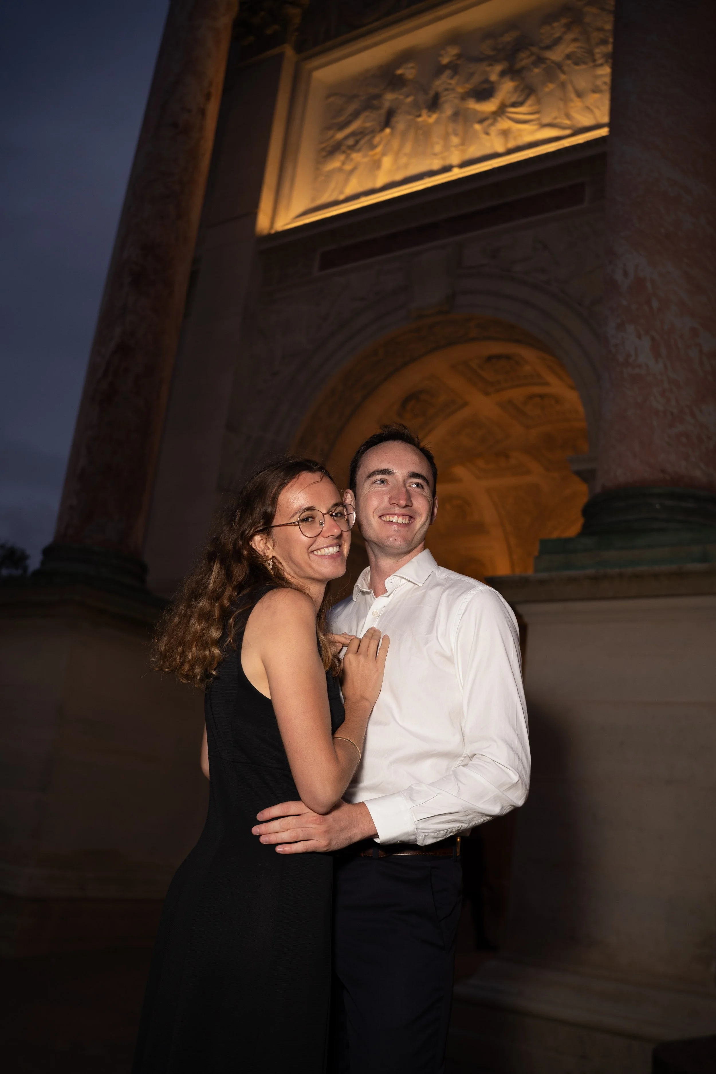 Un couple souriant en soirée devant l'Arc de Triomphe à Paris, la femme porte une robe noire et des lunettes, l'homme porte une chemise blanche.