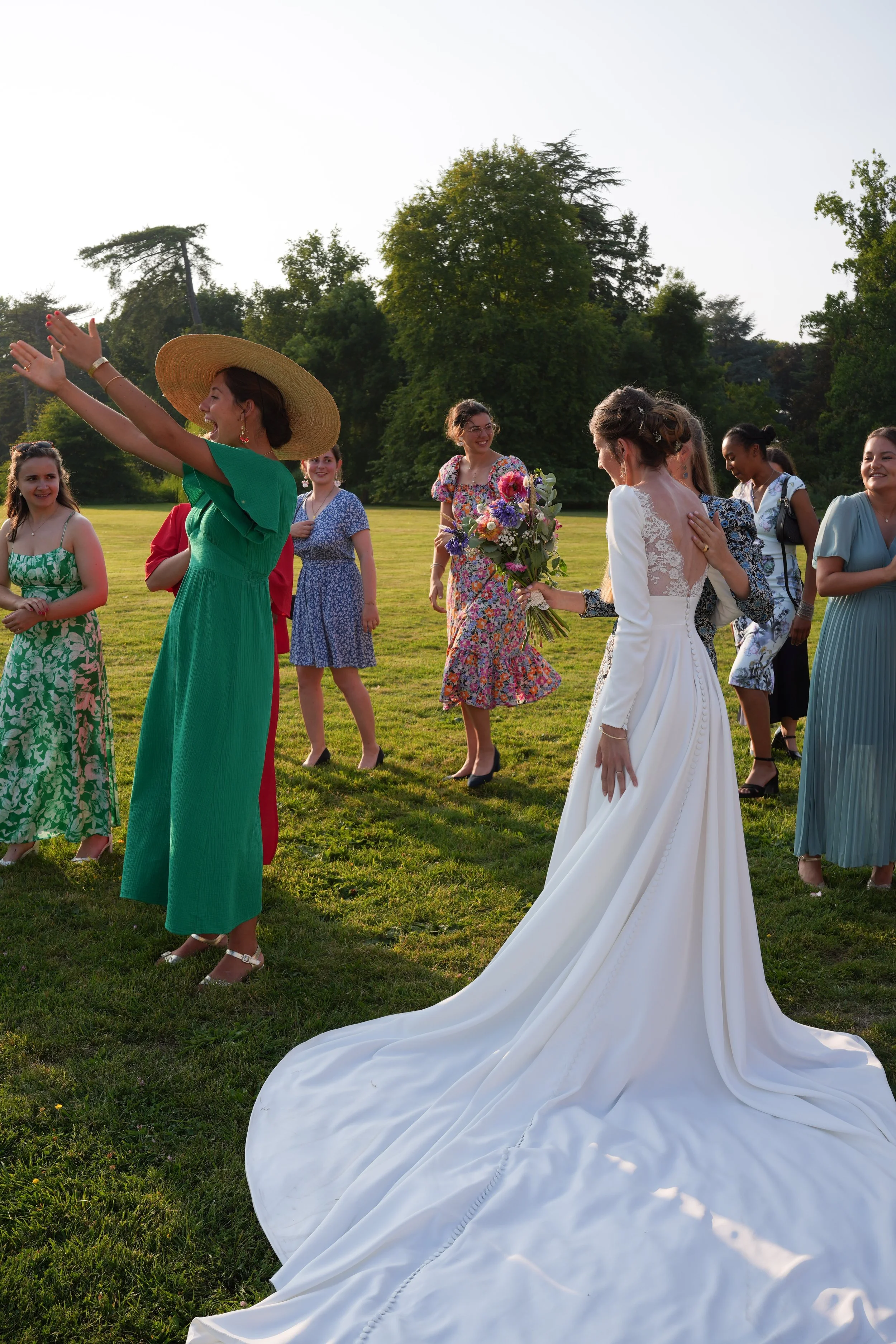 Un groupe de femmes lors d'un mariage en plein air, avec la mariée en robe blanche et une femme avec un grand chapeau discutant et riant, dans un parc vert avec des arbres en arrière-plan.