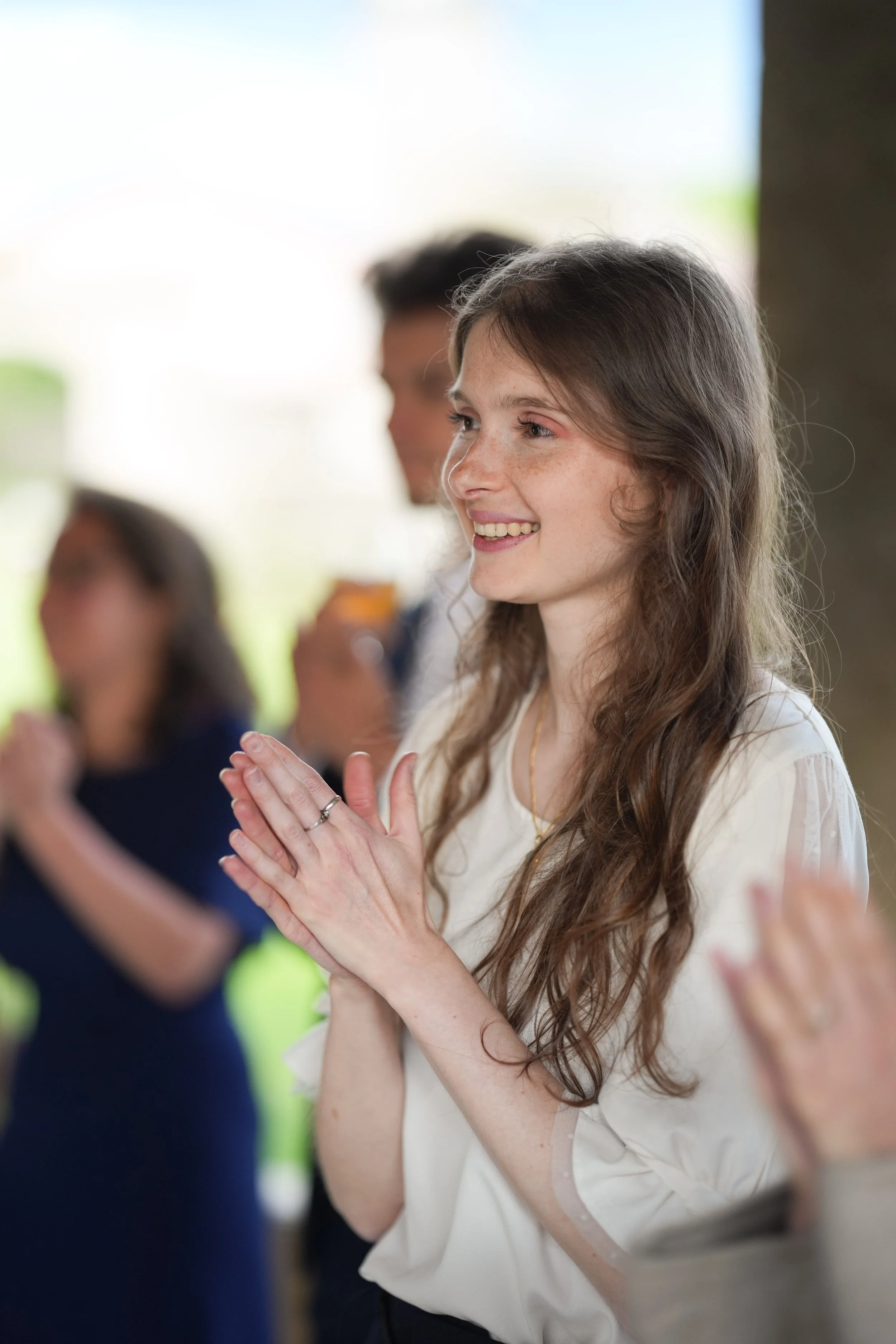 Une jeune femme avec des cheveux longs, bouclés et clairs, souriante et en train de clapper, entourée de plusieurs autres personnes dans un environnement lumineux, probablement à un événement social ou professionnel.
