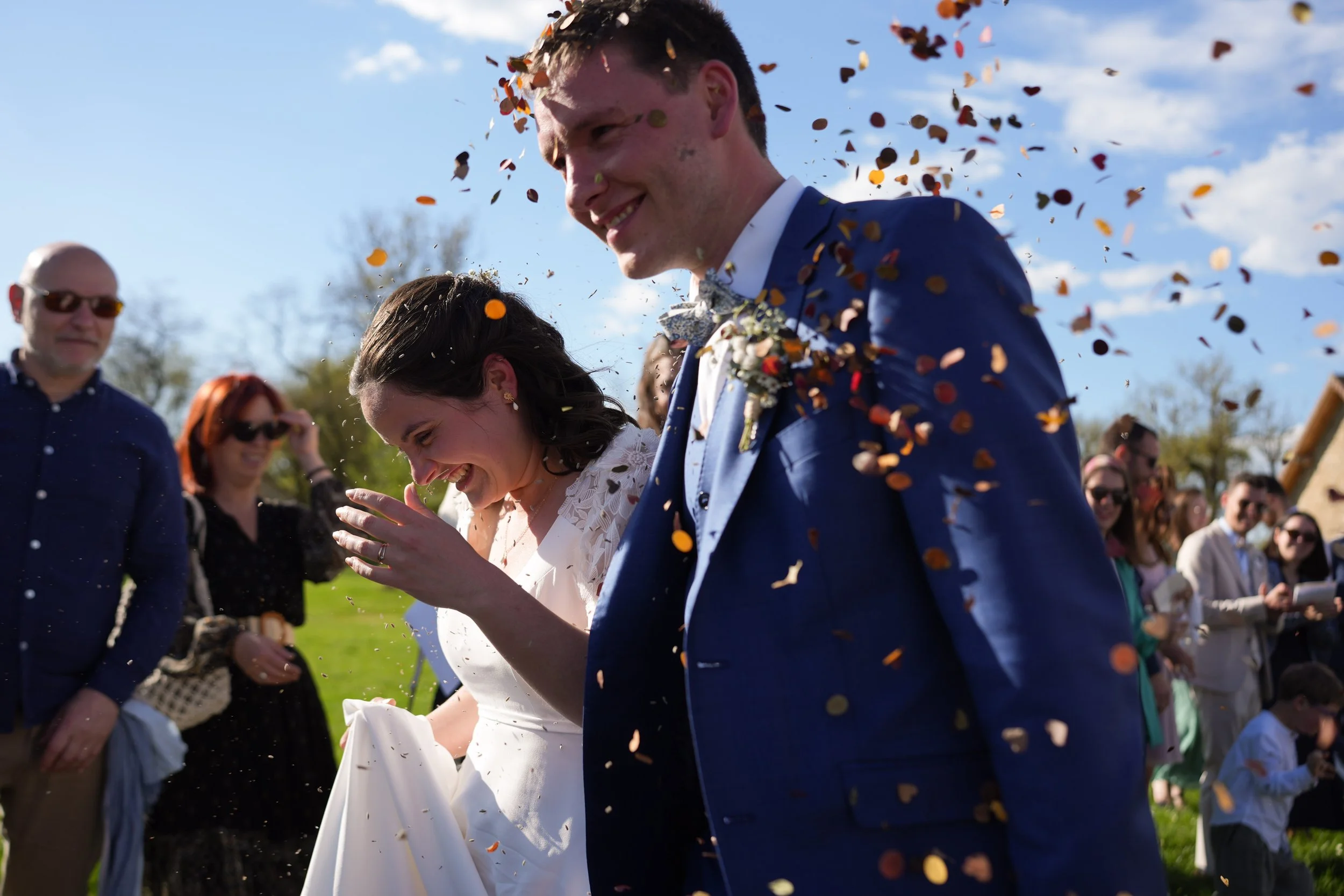 Un couple en costume de mariage, entouré de spectateurs, qui célèbre leur mariage en lançant des confettis en plein air sous un ciel bleu.