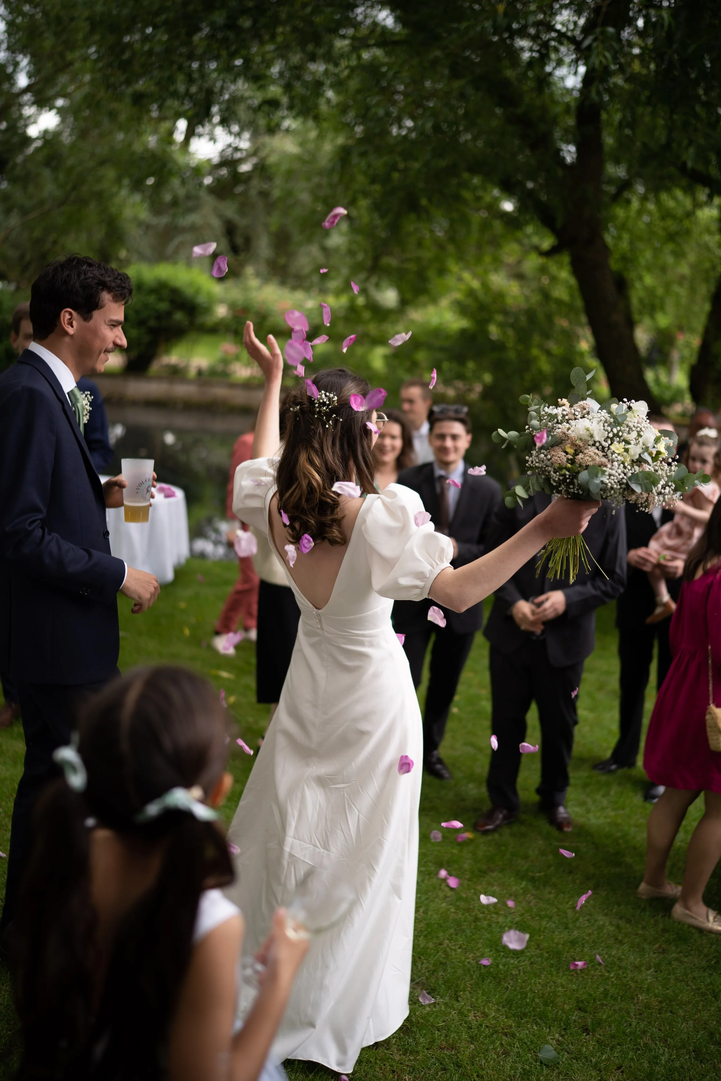 Un groupe de personnes lors d'un mariage, avec une femme en robe blanche tenant un bouquet de fleurs, entourée de amis et famille, dans un parc avec des arbres
