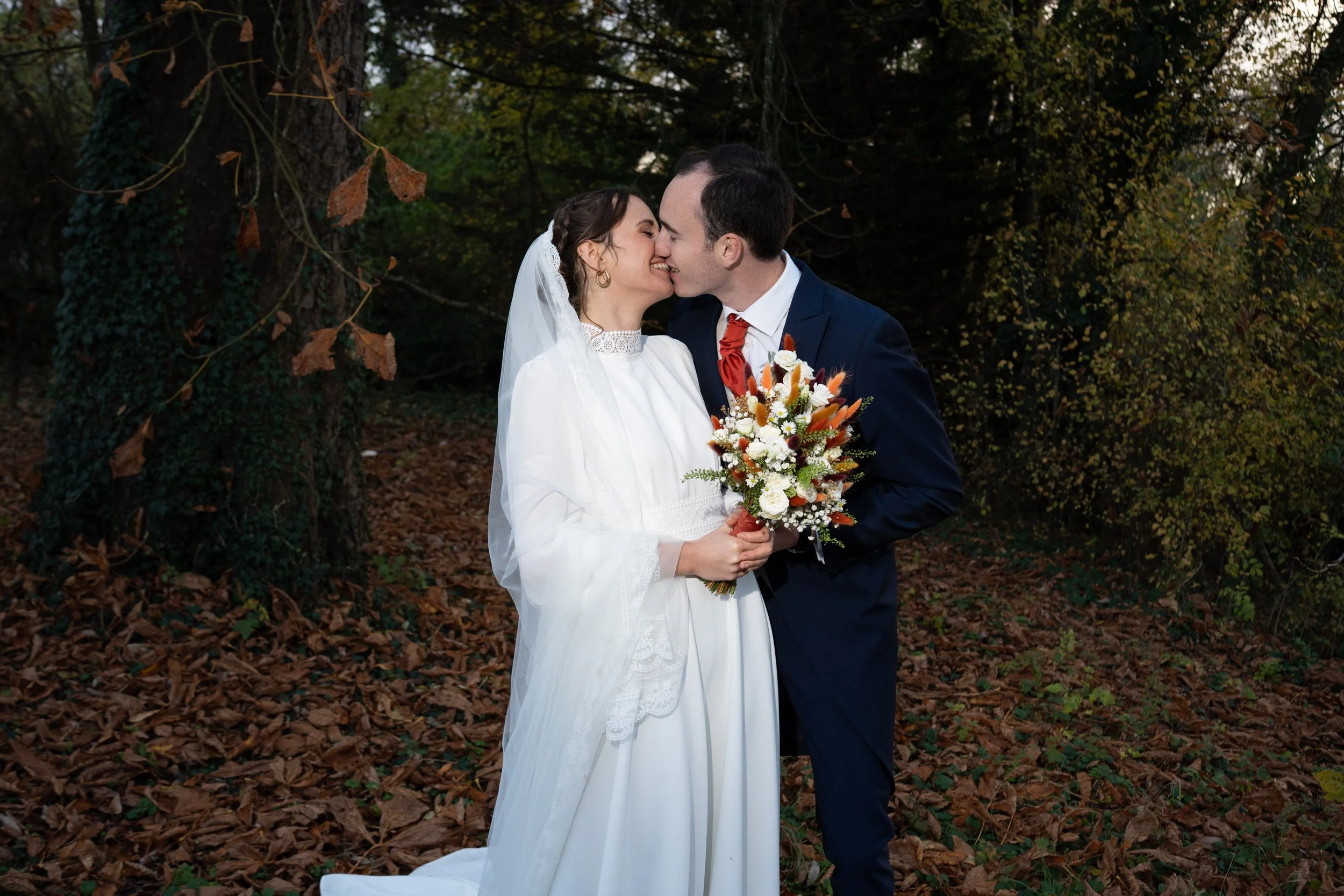 Un couple de mariés s'embrassant dans une forêt, la femme portant une robe blanche et un voile, tenant un bouquet de fleurs, et l'homme en costume bleu avec une cravate rouge.