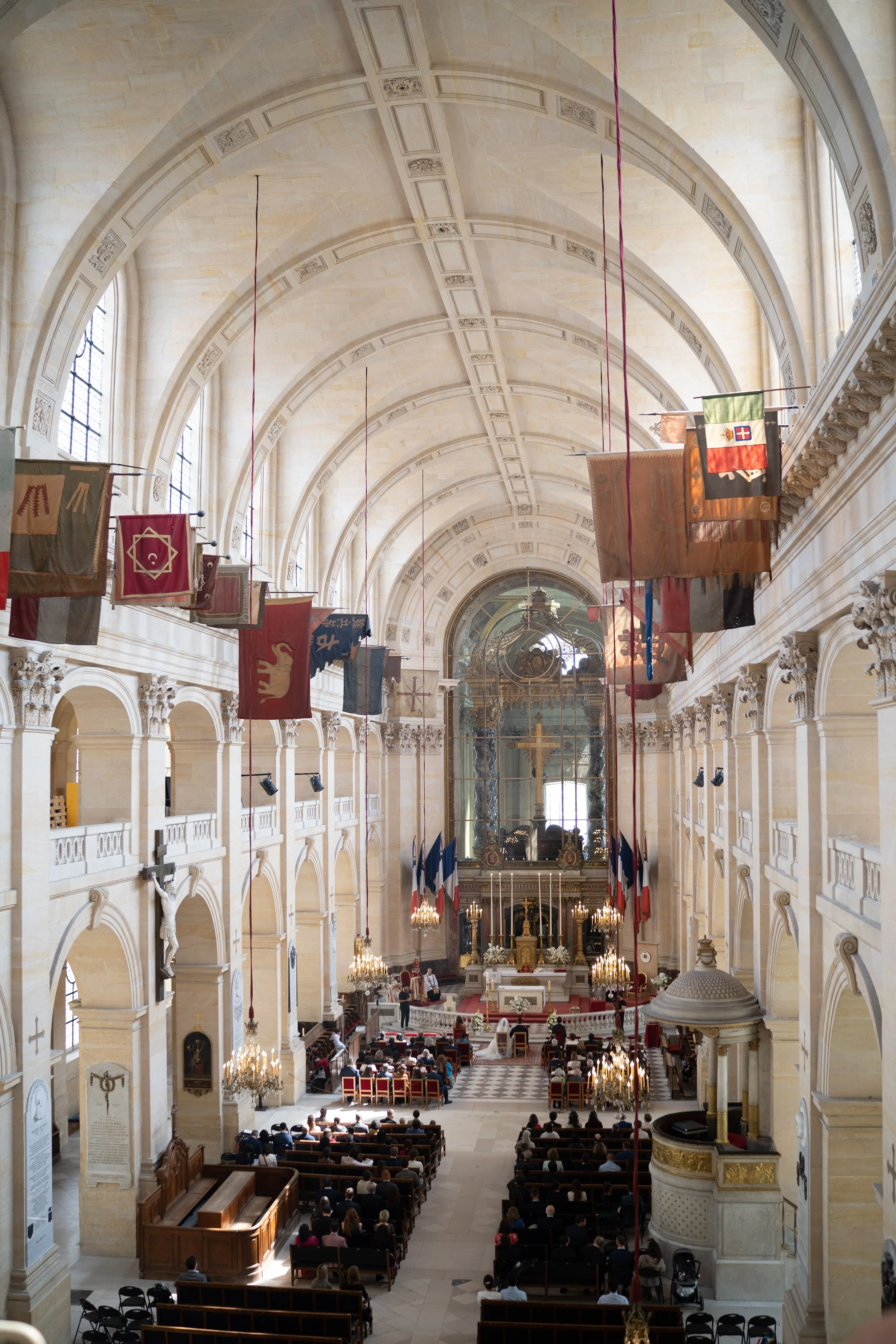 Intérieur d'une grande église ou cathédrale avec des arches hautes, des drapeaux suspendus, des chandeliers et des personnes assises pour une cérémonie ou une messe.