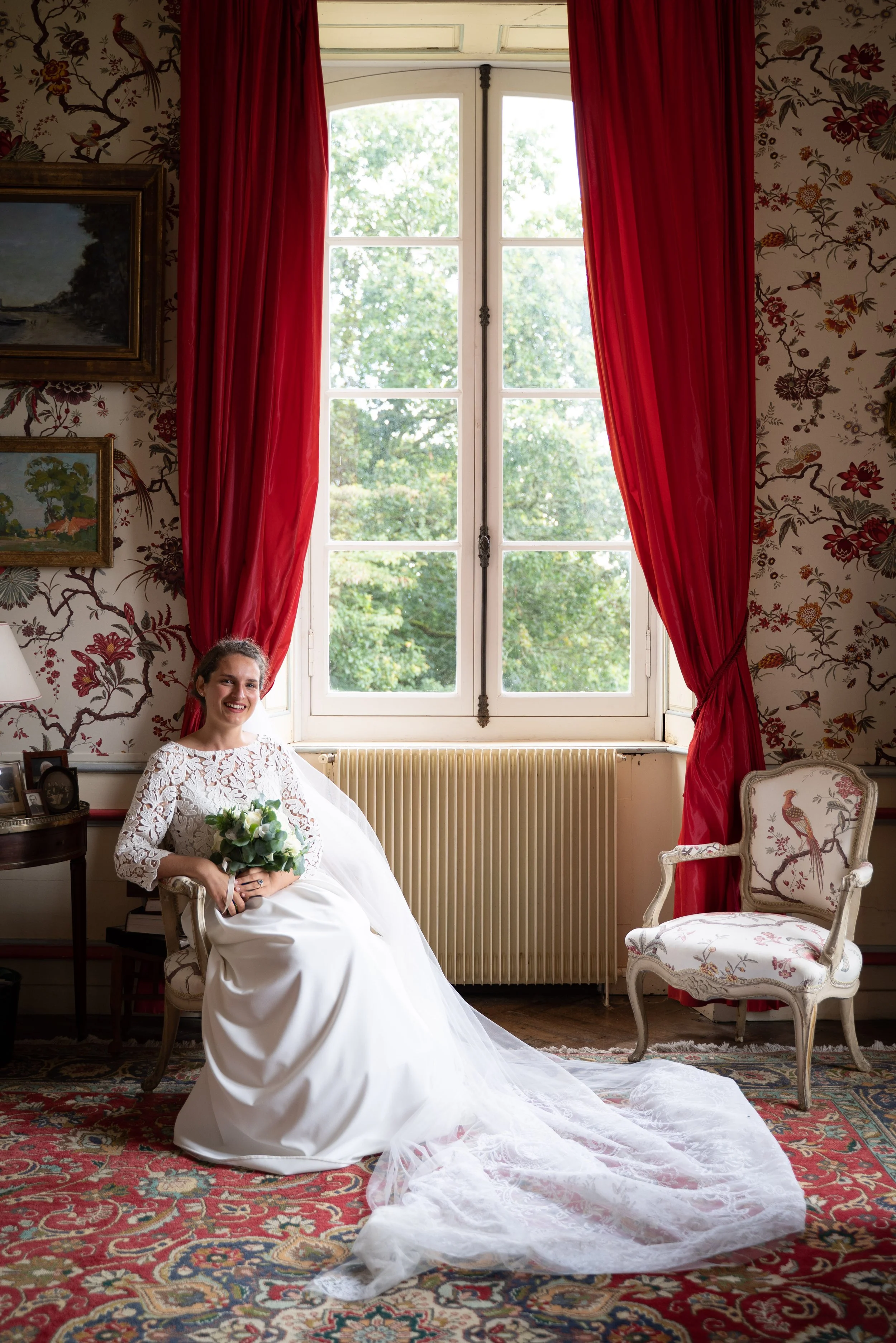 Une femme souriante habillée d'une robe de mariage blanche, assise dans une pièce ornée de rideaux rouges, de tapis coloré, de papier peint floral, avec une grande fenêtre laissant entrer la lumière naturelle, et entourée de meubles anciens et de tab