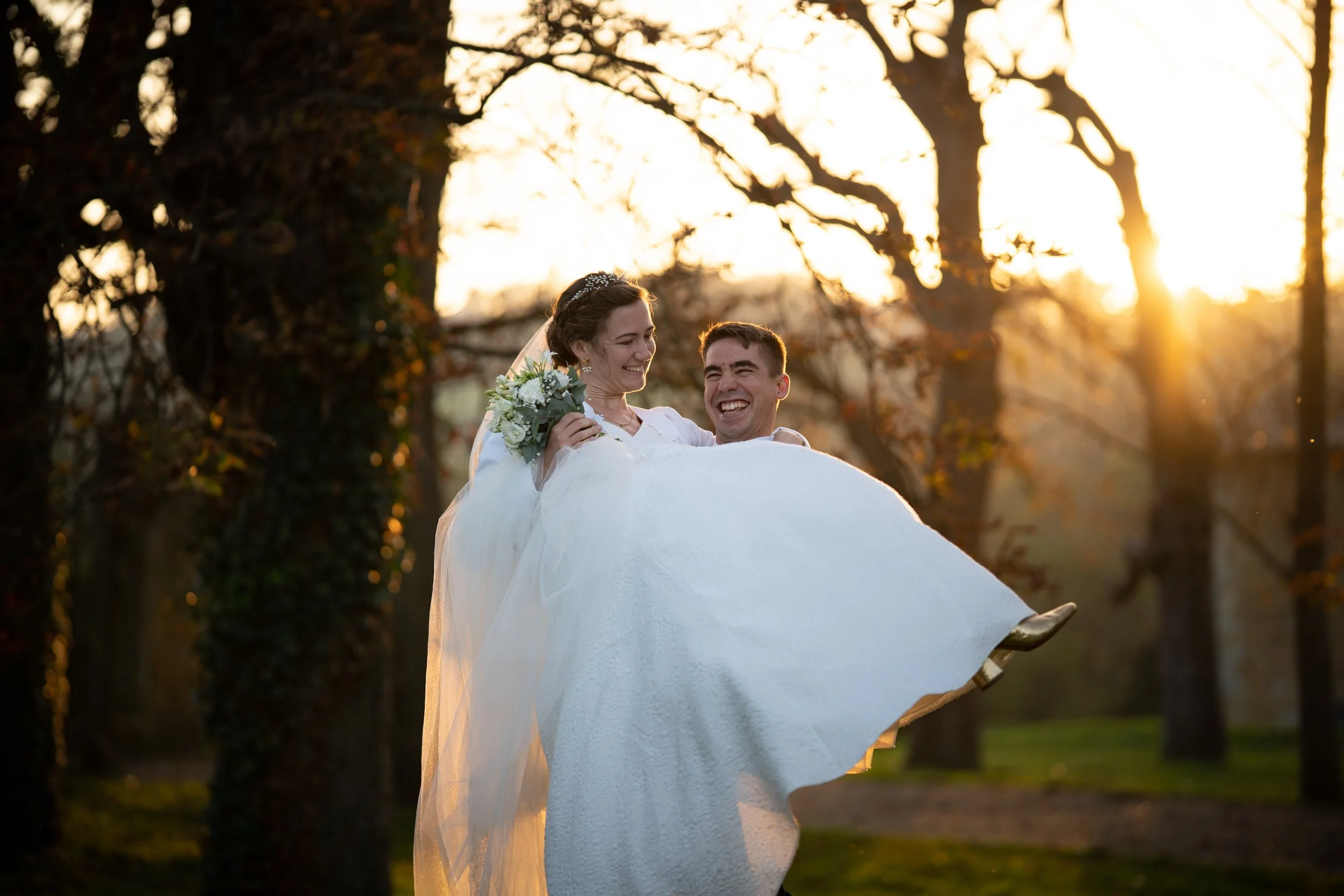 Un couple de mariés souriants, portant une robe de mariée blanche et un costume, est porté par un homme dans un décor extérieur au coucher du soleil avec des arbres en arrière-plan.