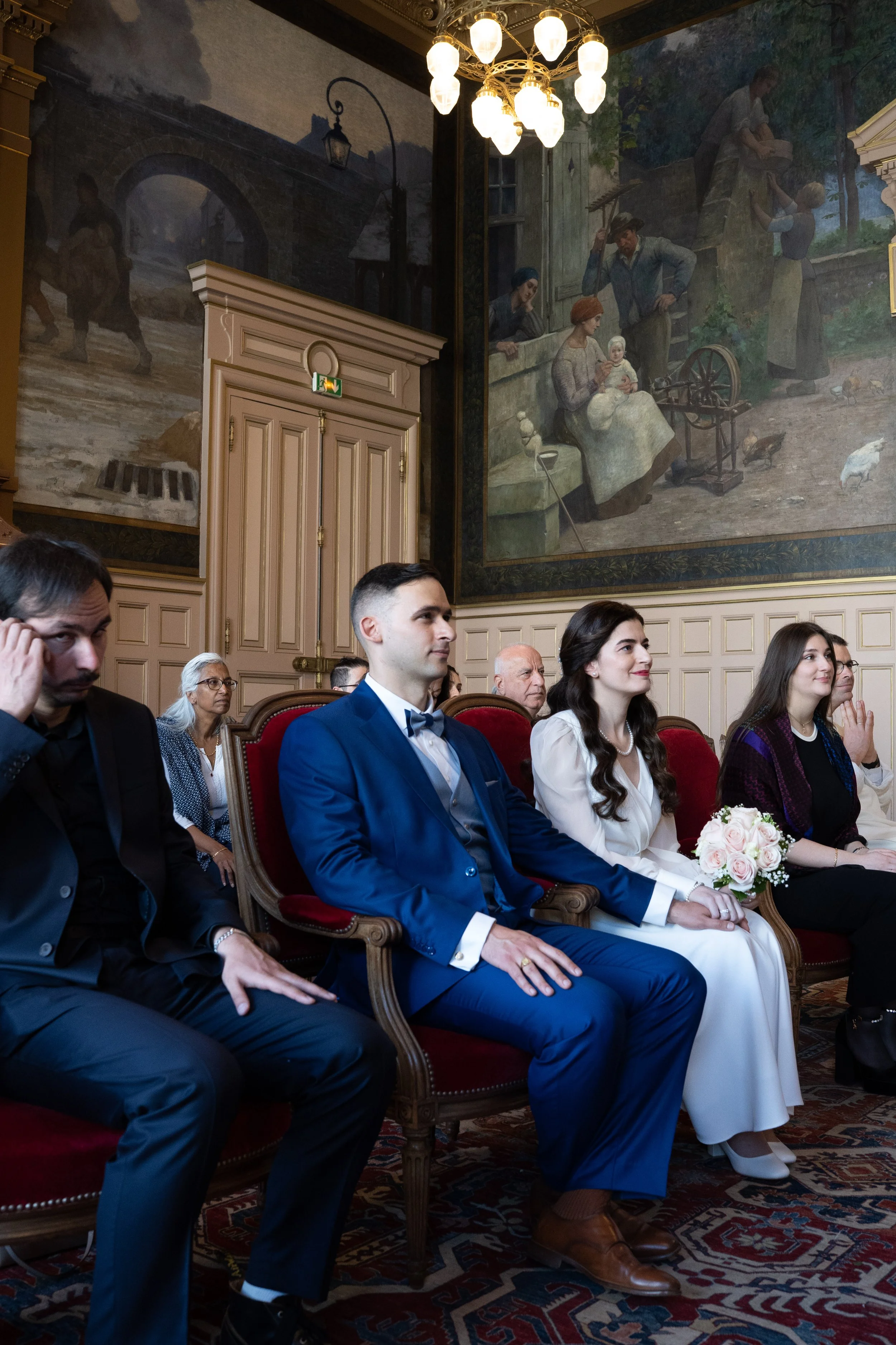 Une scène d'un mariage avec des personnes assises, dont un couple en costume et robe blanche au premier plan, dans une salle ornée de grands tableaux.