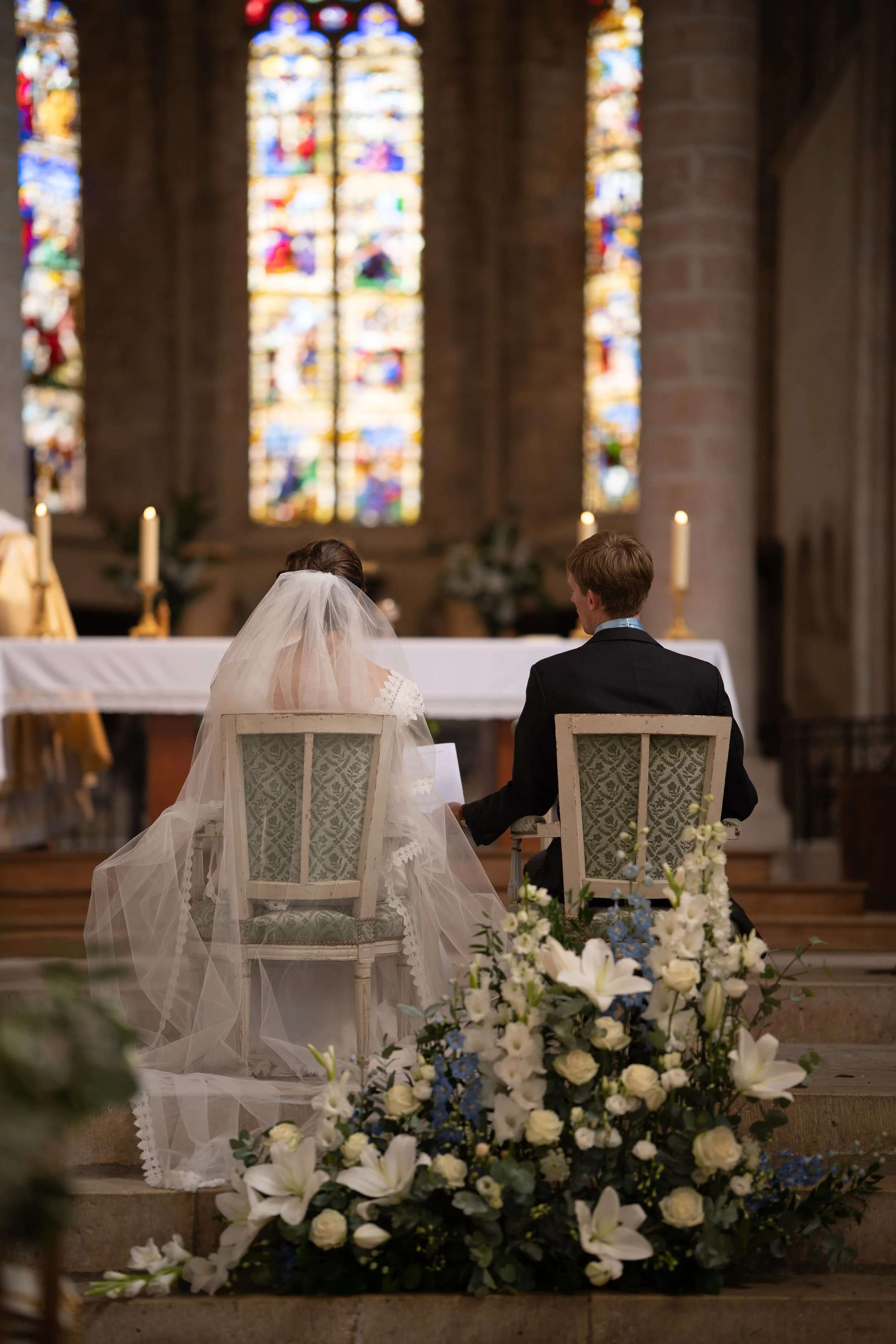 Un couple assis sur des chaises dans une église lors d'une cérémonie de mariage, entouré de fleurs blanches et de lys, avec des vitraux colorés en arrière-plan.