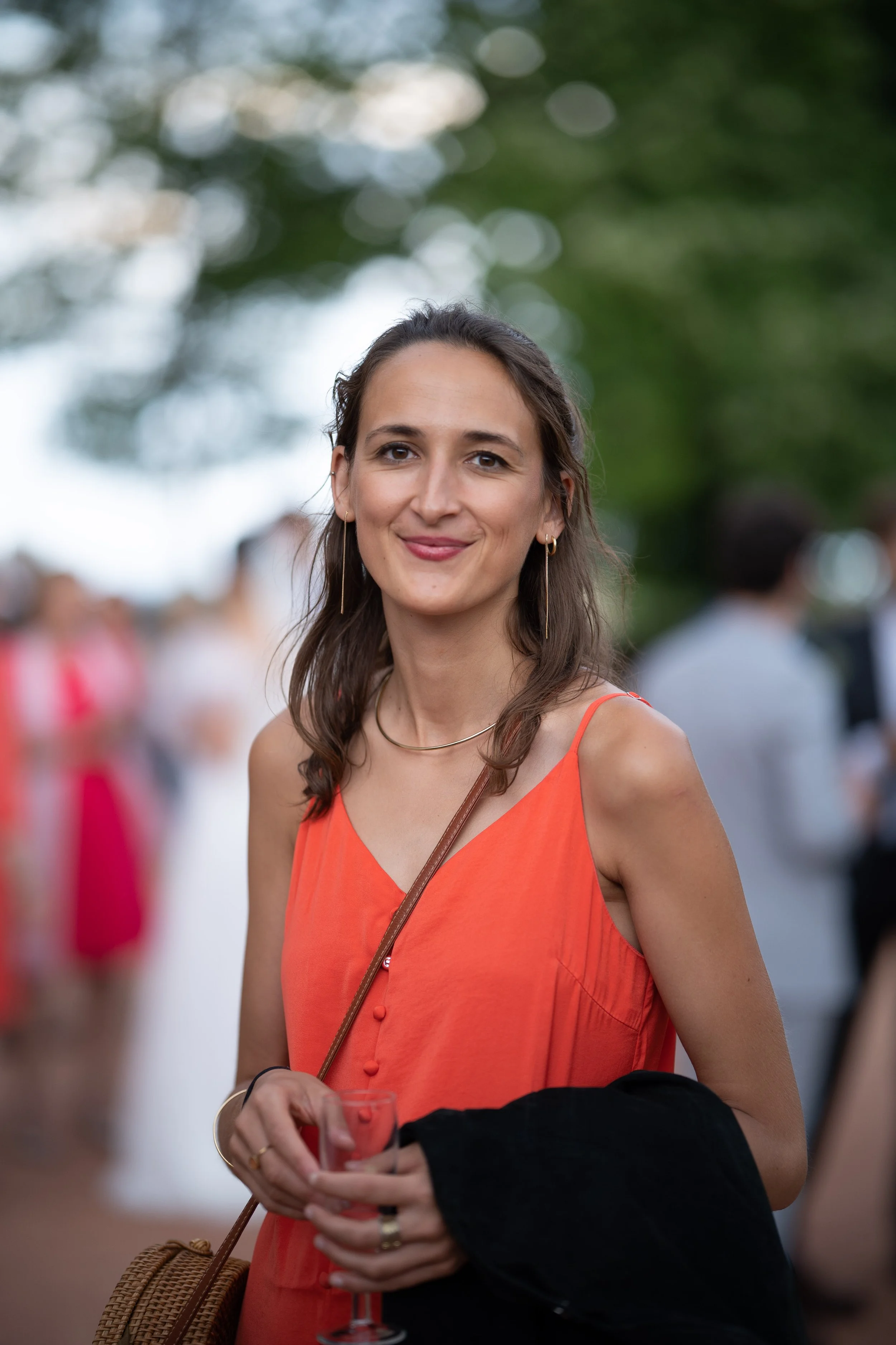 Une femme souriante portant une robe orange, tenant un verre à vin lors d'une réunion en plein air, avec d'autres personnes floues en arrière-plan.