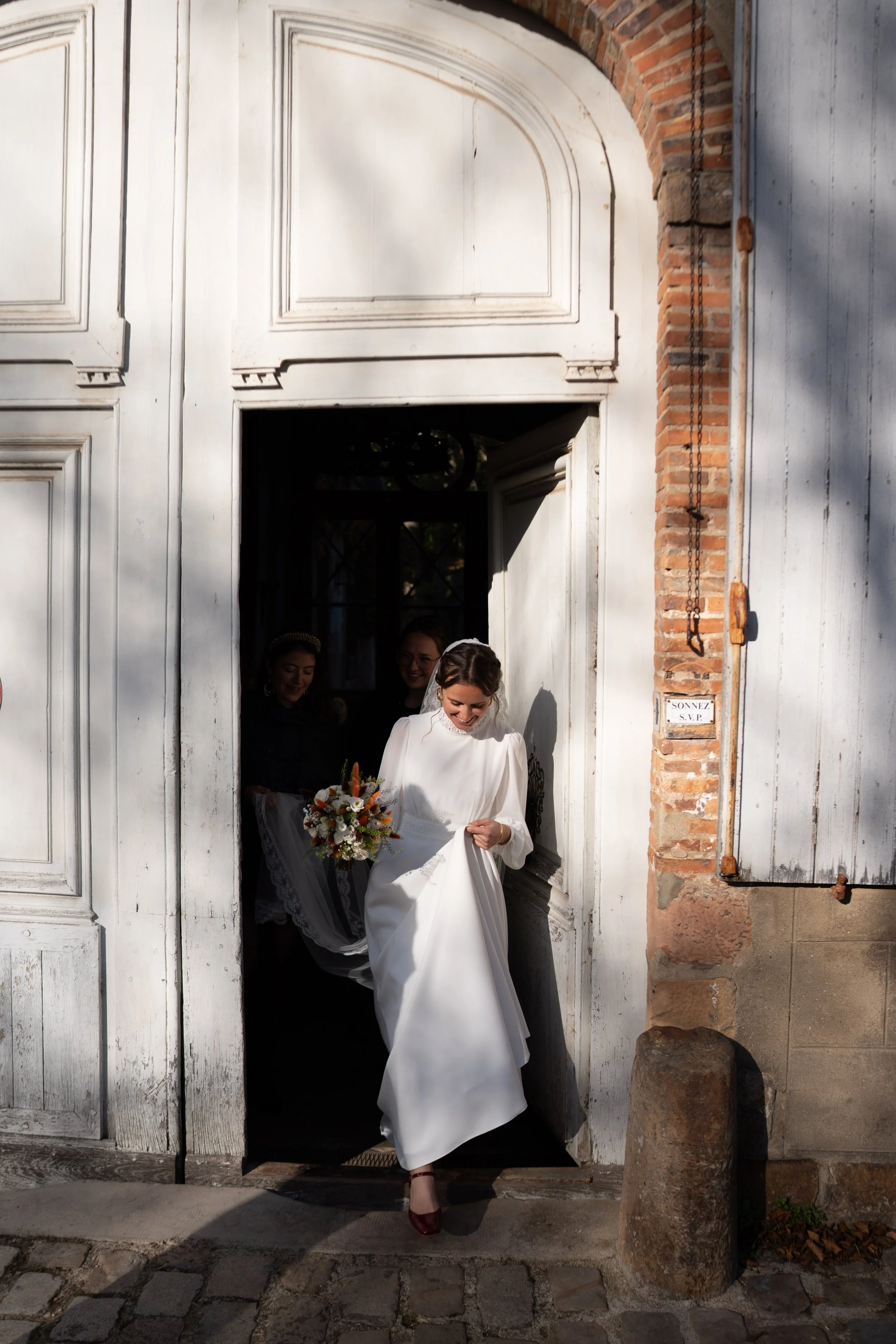Une femme en robe de mariage sort d'une vieille porte en bois blanche, tenant un bouquet de fleurs. Deux autres femmes derrière l'elle l'accompagnent. La porte est encadrée par un mur en briques. La scène semble se passer à l'extérieur, à proximité d'une vieille bâtisse.