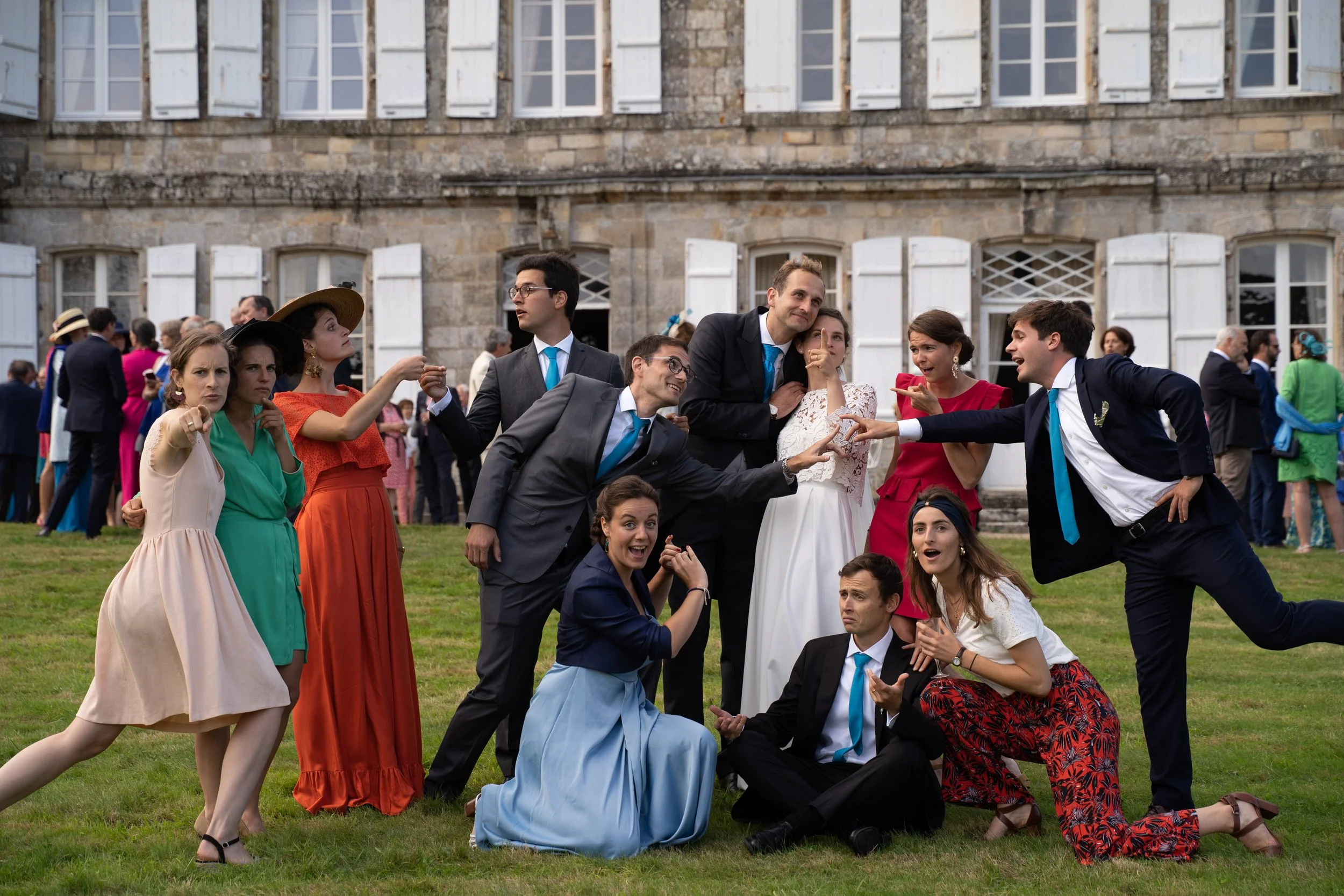 Groupe de personnes habillées de façon élégante, posant et interagissant devant un bâtiment en pierre avec des fenêtres à volets blancs. Il semble s'agir d'une photo prise lors d'une célébration ou d'un événement social en plein air.