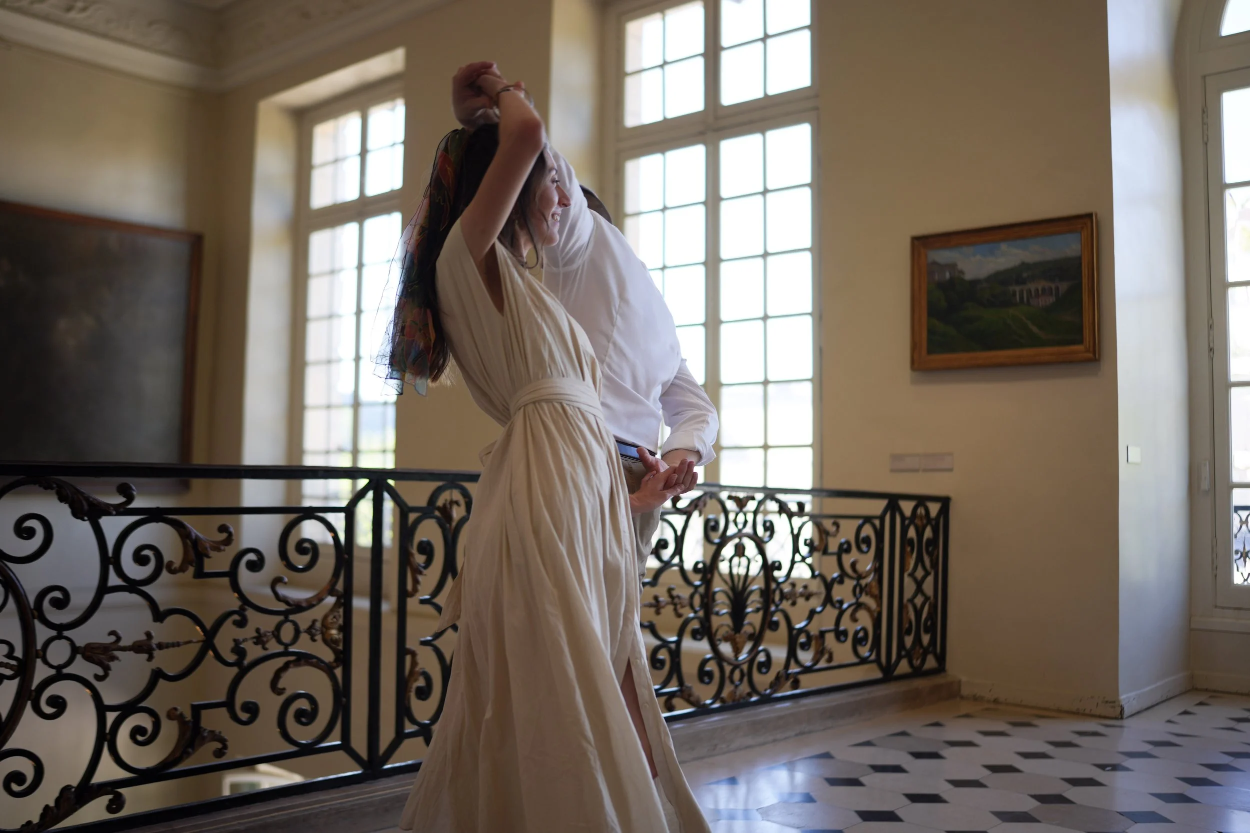 Une femme souriante portant une robe beige, debout près d'une rampe en fer forgé dans une salle lumineuse avec de grandes fenêtres, une peinture dans un cadre en bois accroché au mur.