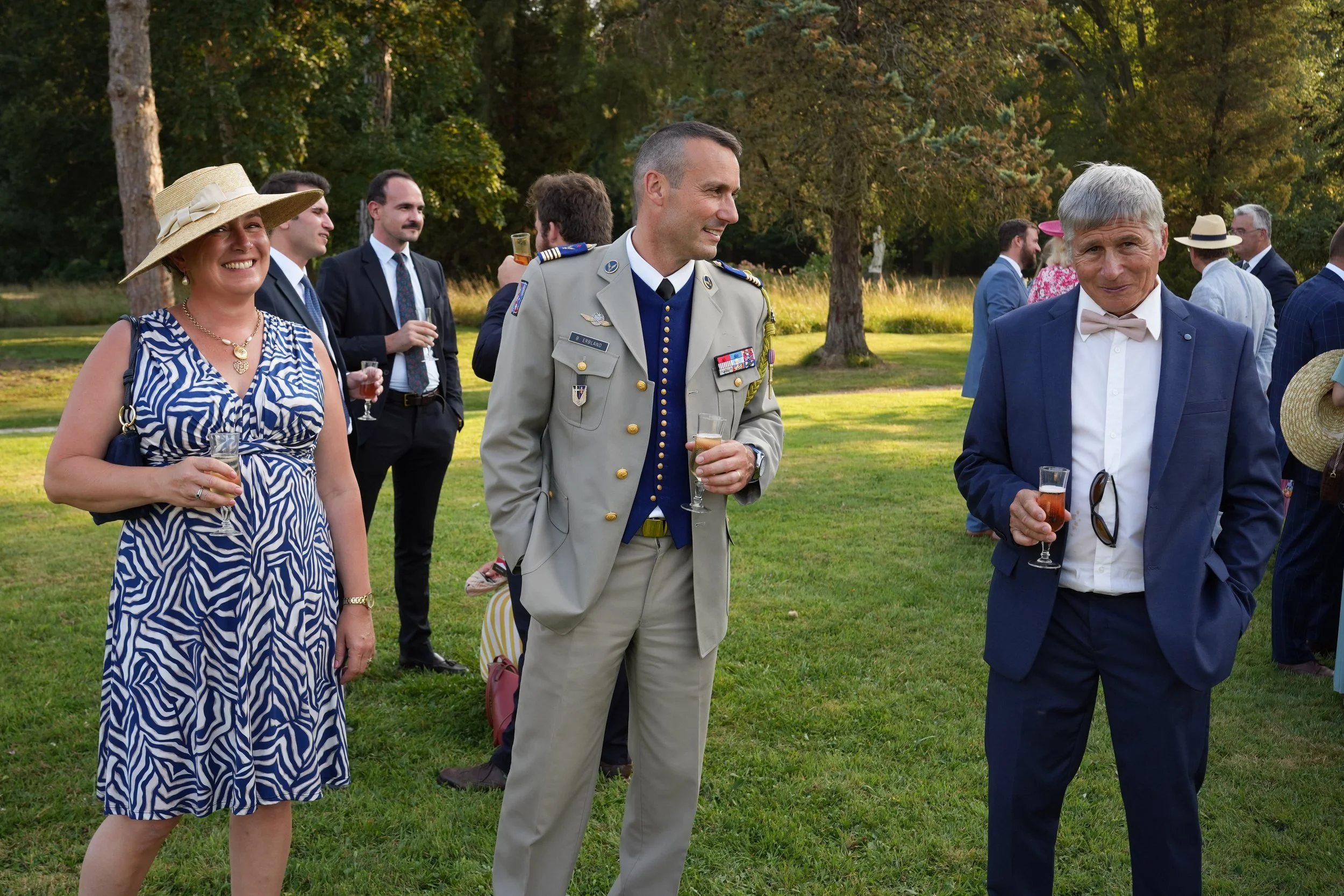 Groupe de personnes habillées élégamment lors d'une fête en plein air, avec des arbres en arrière-plan.