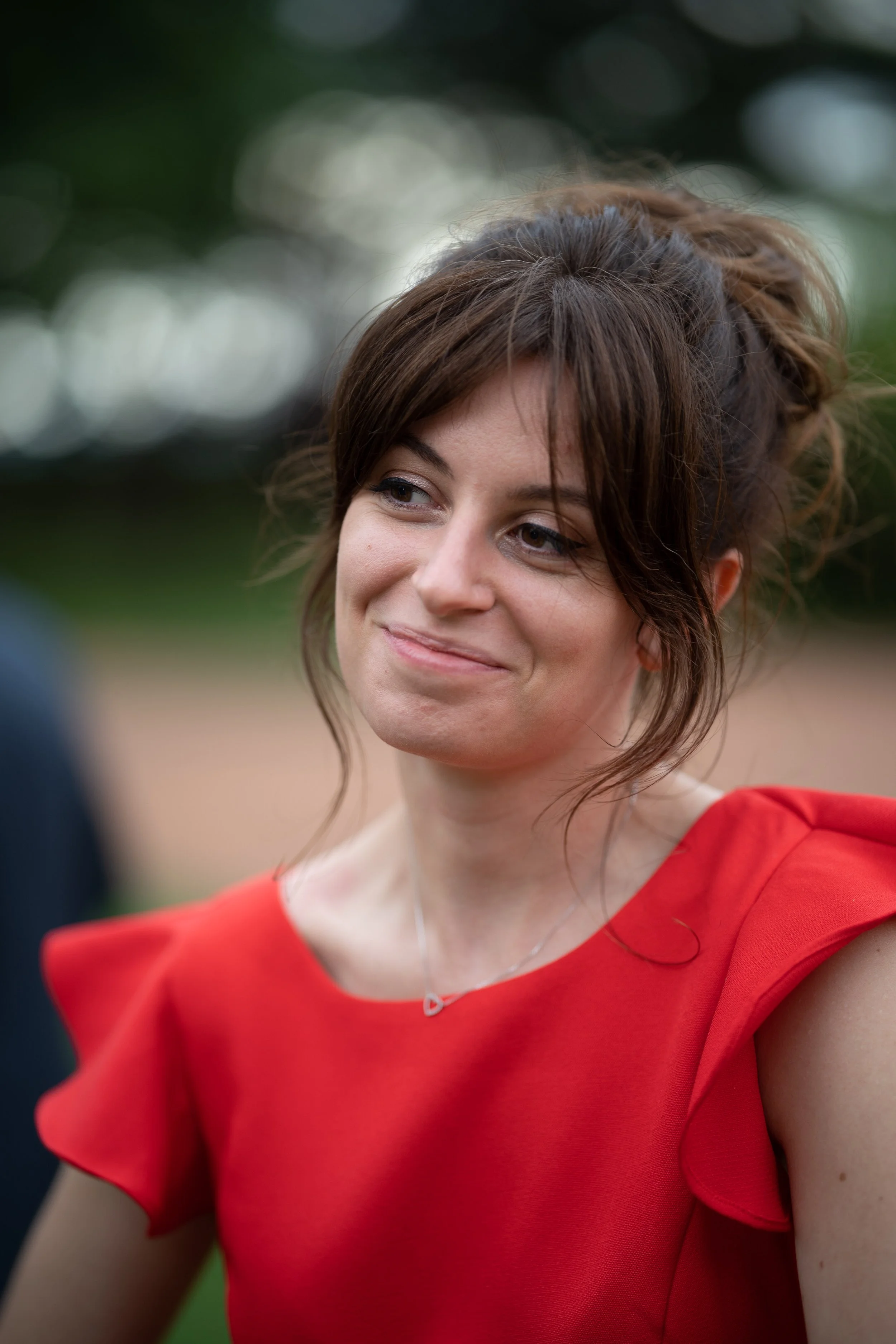 Une femme avec des cheveux bruns attachés en chignon, portant une robe rouge à manches courtes, souriant légèrement avec un fond flou de verdure.